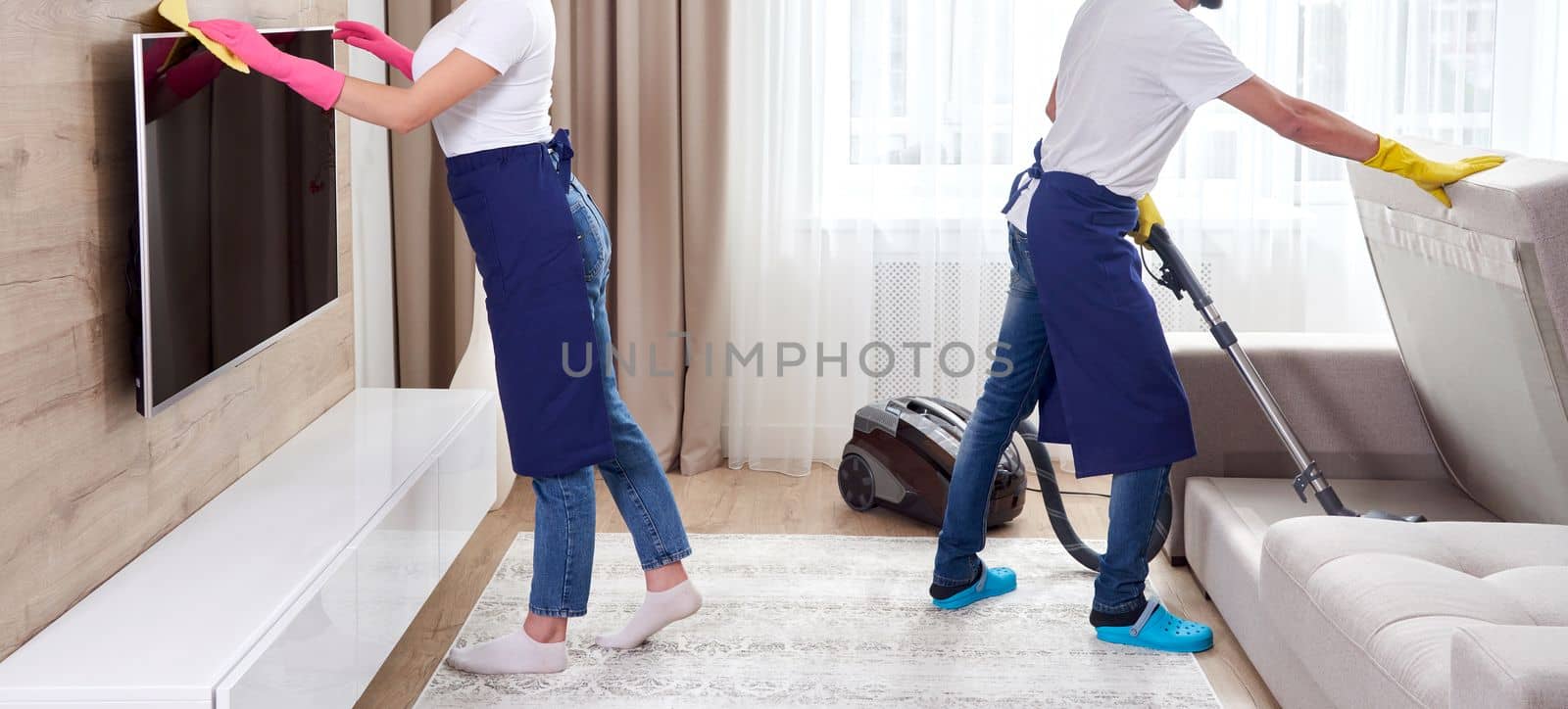 Professional cleaners in blue uniform washing floor and wiping dust from the furniture in the living room of the apartment. Cleaning service concept by Mariakray