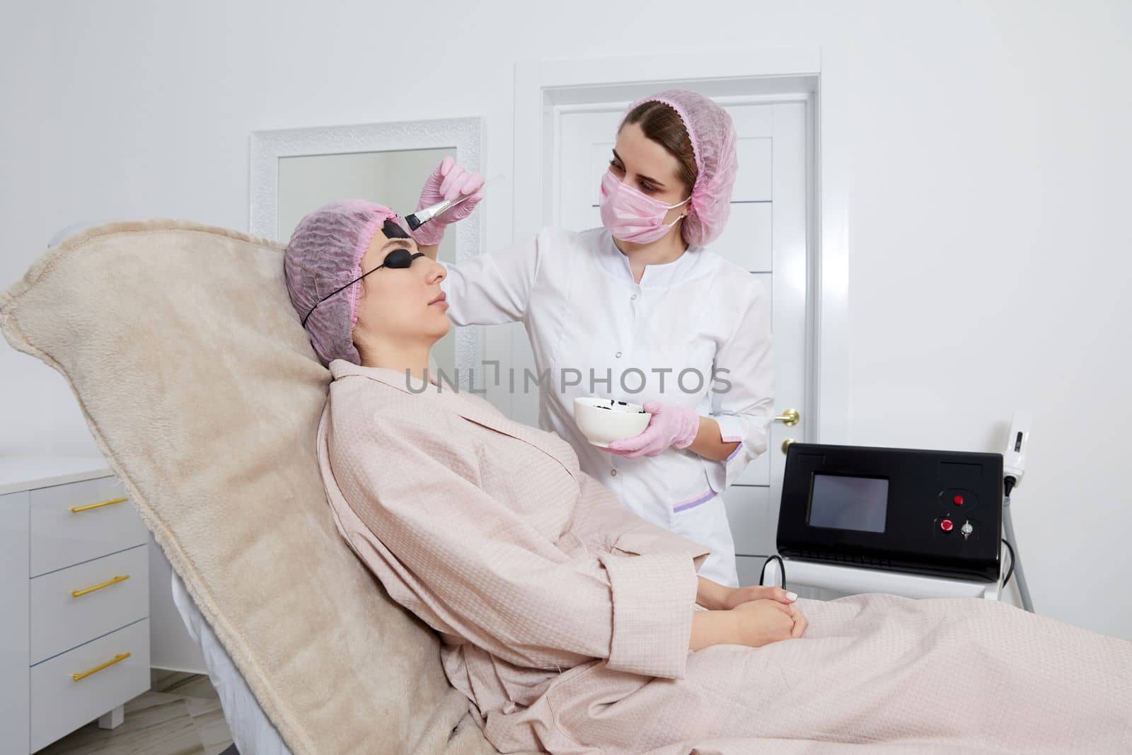 Cosmetologist applying black mask on the face of a beautiful woman for carbon peeling in beauty salon