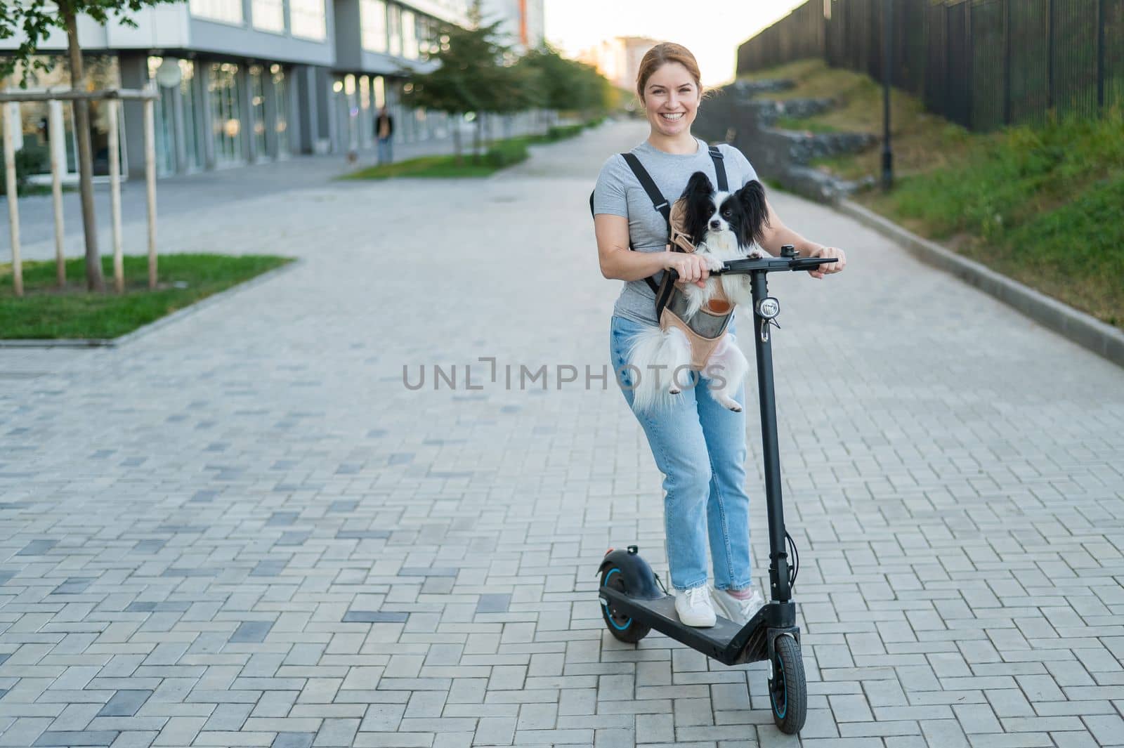 A woman rides an electric scooter with a dog in a backpack. Pappilion Spaniel Continental in a sling