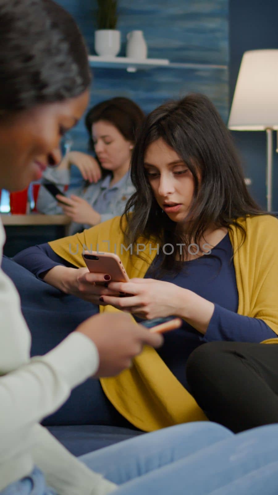 Group of mixed race people hanging out together late at night in living room searching online news on mobile. Multiracial friends watching entertainment movie during surprise party