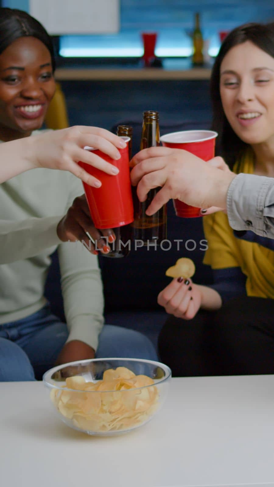 Multi-cultural friends socializing while sitting on sofa in living room late at night drinking beer enjoying home party. Group of mixed race people hanging out enjoying time spending together