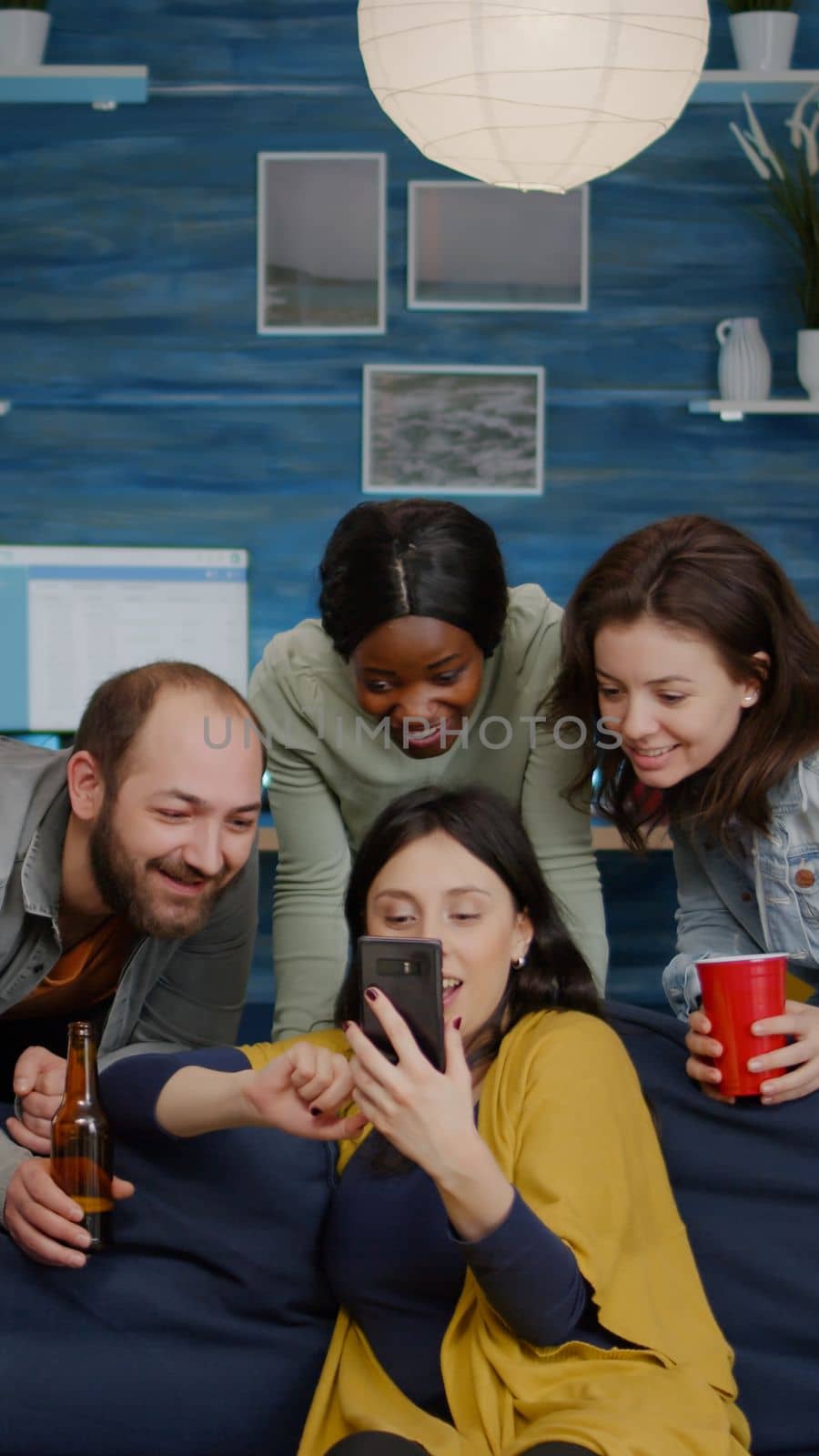 Multiracial friends hanging out together socializing while looking at funny video on smartphone. Group of multi-ethnic people sitting on sofa late at night in living room drinking beer