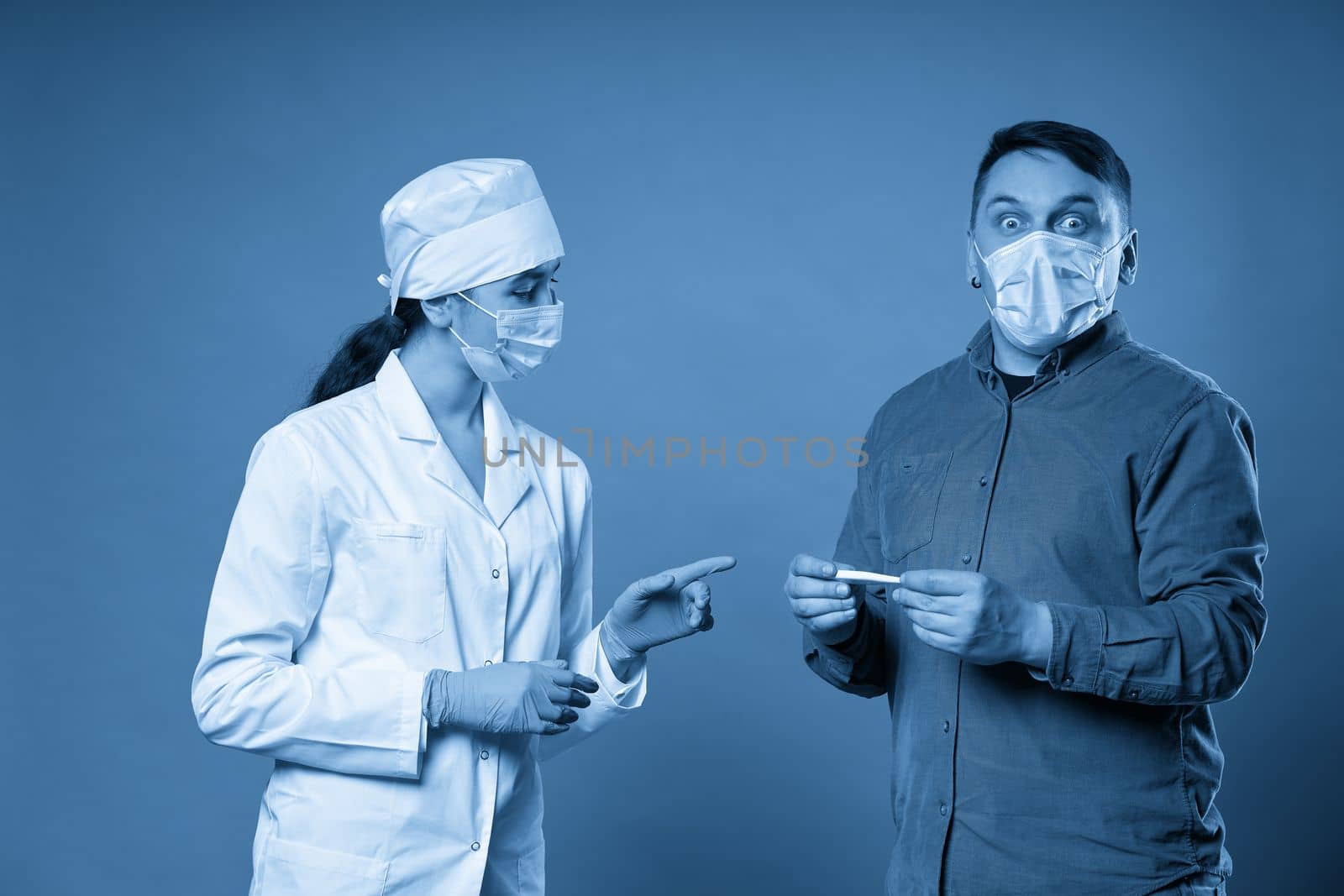 Young doctor measuring temperature of a patient with thermometer, studio shot on blue background by Mariakray