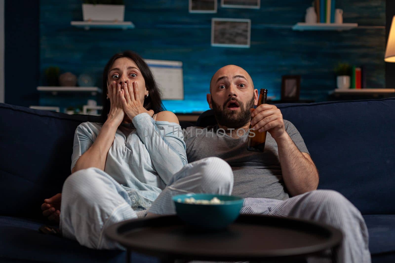 POV of afraid couple watching scary movie on television program, drinking beer and eating popcorn. Man and woman feeling shocked, surprised and terrified looking at horror film on TV.