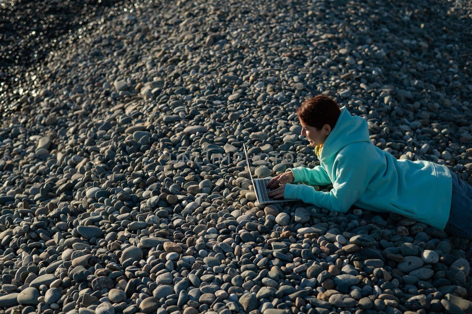 Caucasian woman lying on her stomach on a pebble beach and typing on a laptop. Remote work freelancer