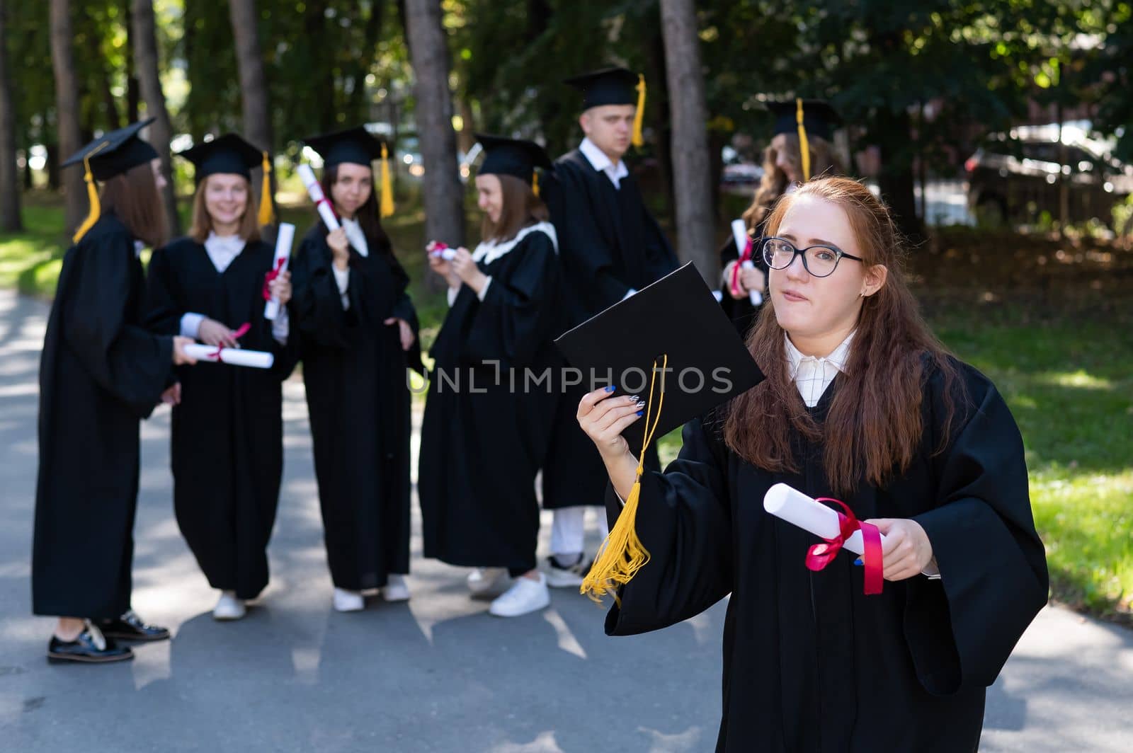 Portrait of a young caucasian woman in glasses and a graduate gown against the background of classmates. A group of graduate students outdoors. by mrwed54