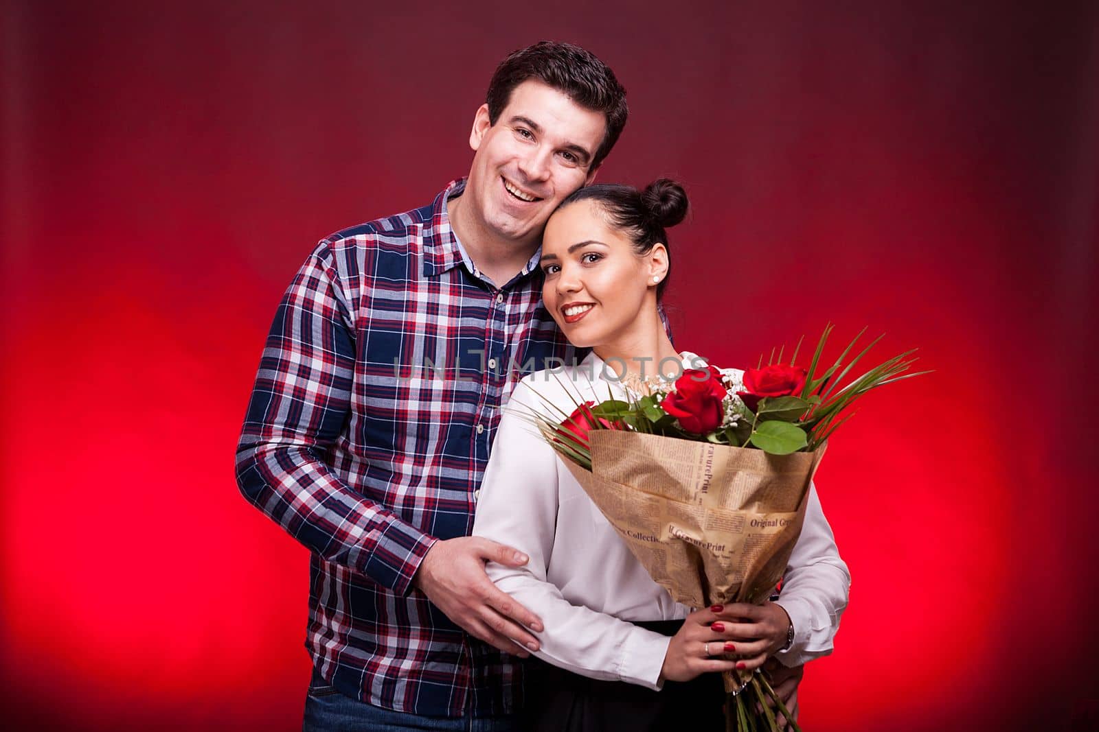 Man embracing her beautiful wife while she holds a roses bouquet in hands on red background in studio photo