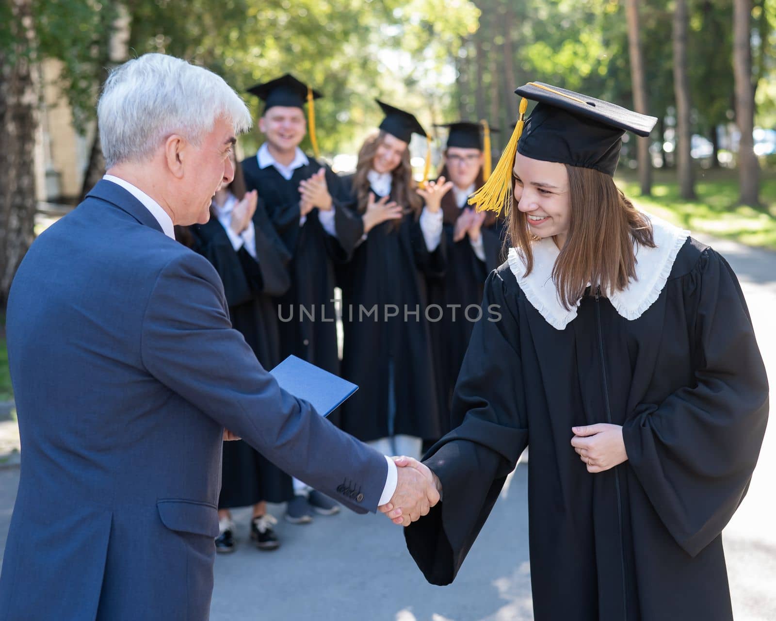 The teacher shakes hands with the student and presents the diploma outdoors. A group of university graduates