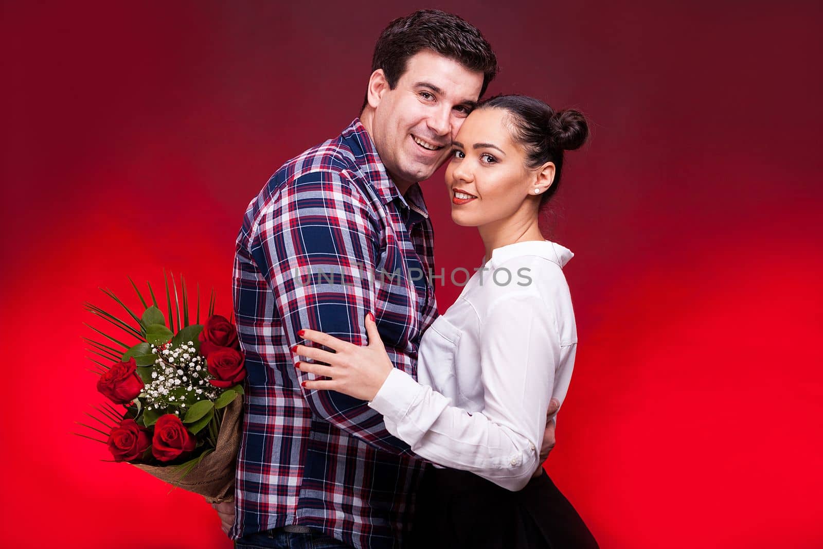 Man with a roses bouquet at his back on a first date embracing a gorgeous girl. Red background and studio photo