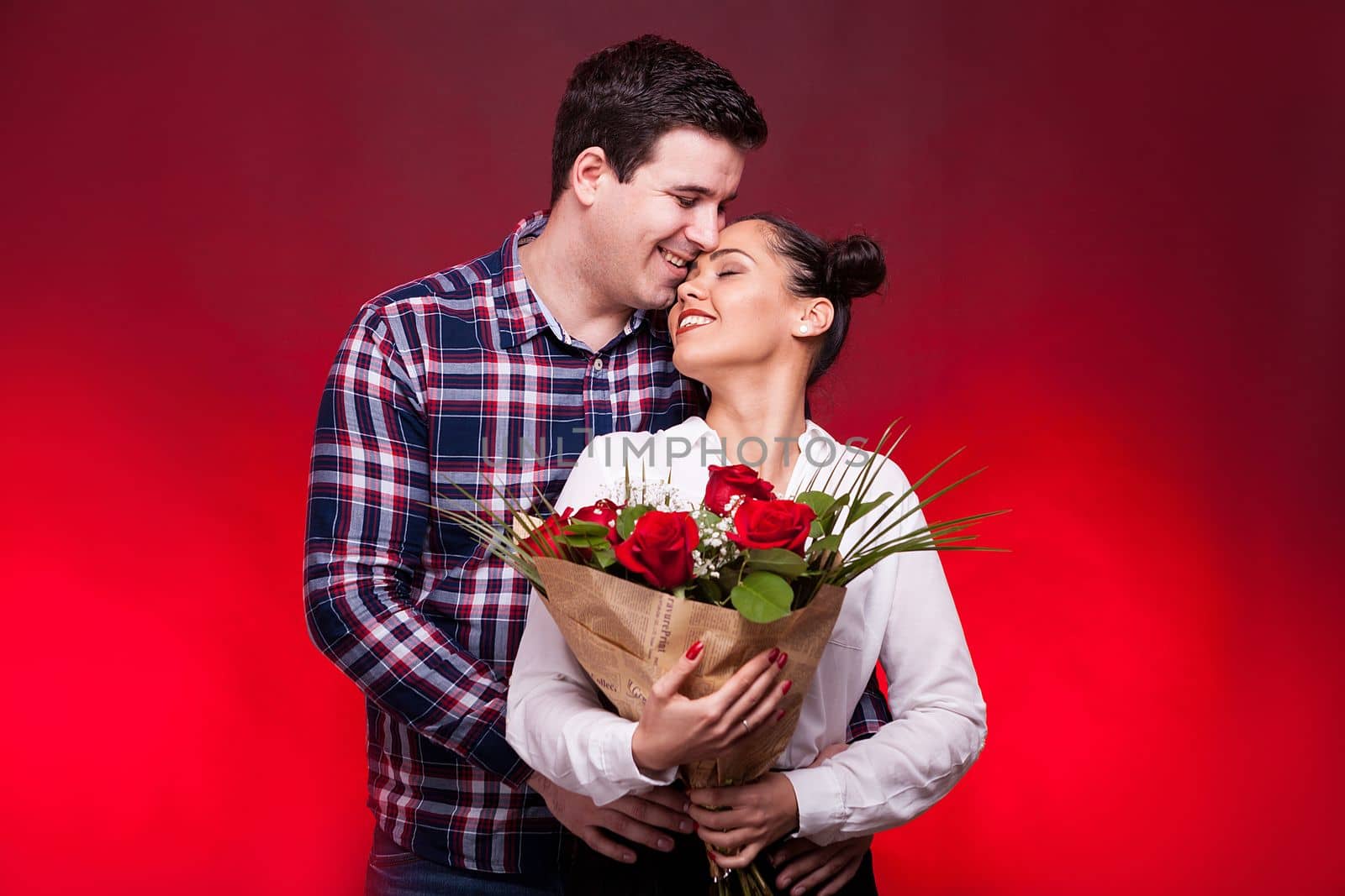 Man embracing her wife and kissing her on the forehead while she holds a roses bouquet in hands on red background in studio photo