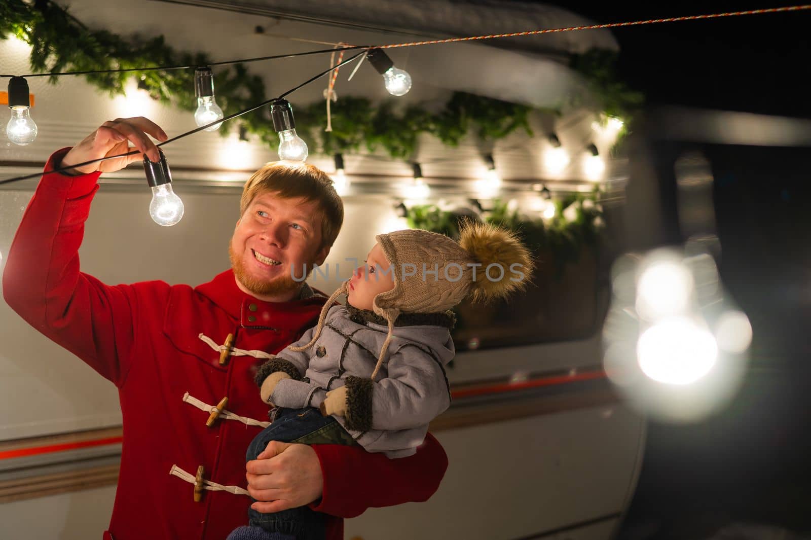 Caucasian red-haired man with a boy in his arms at the mobile home. Father and son celebrate Christmas on a trip