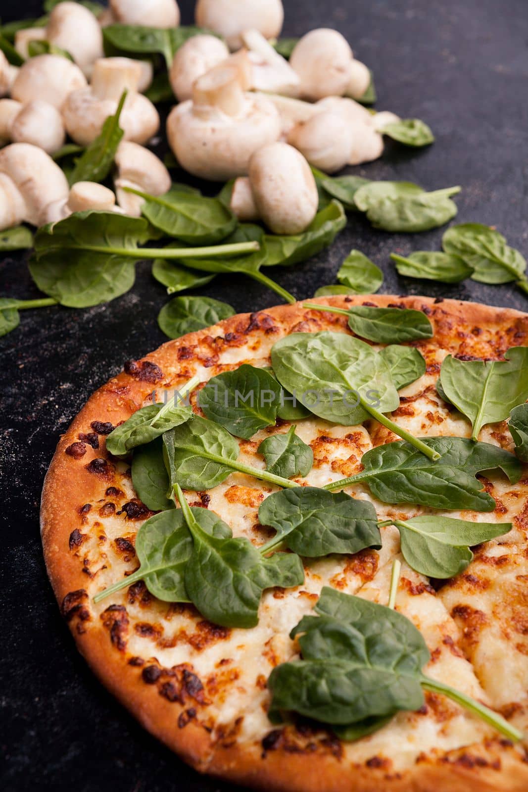Pizza covered with spinach leaves next to mushrooms on dark wooden background