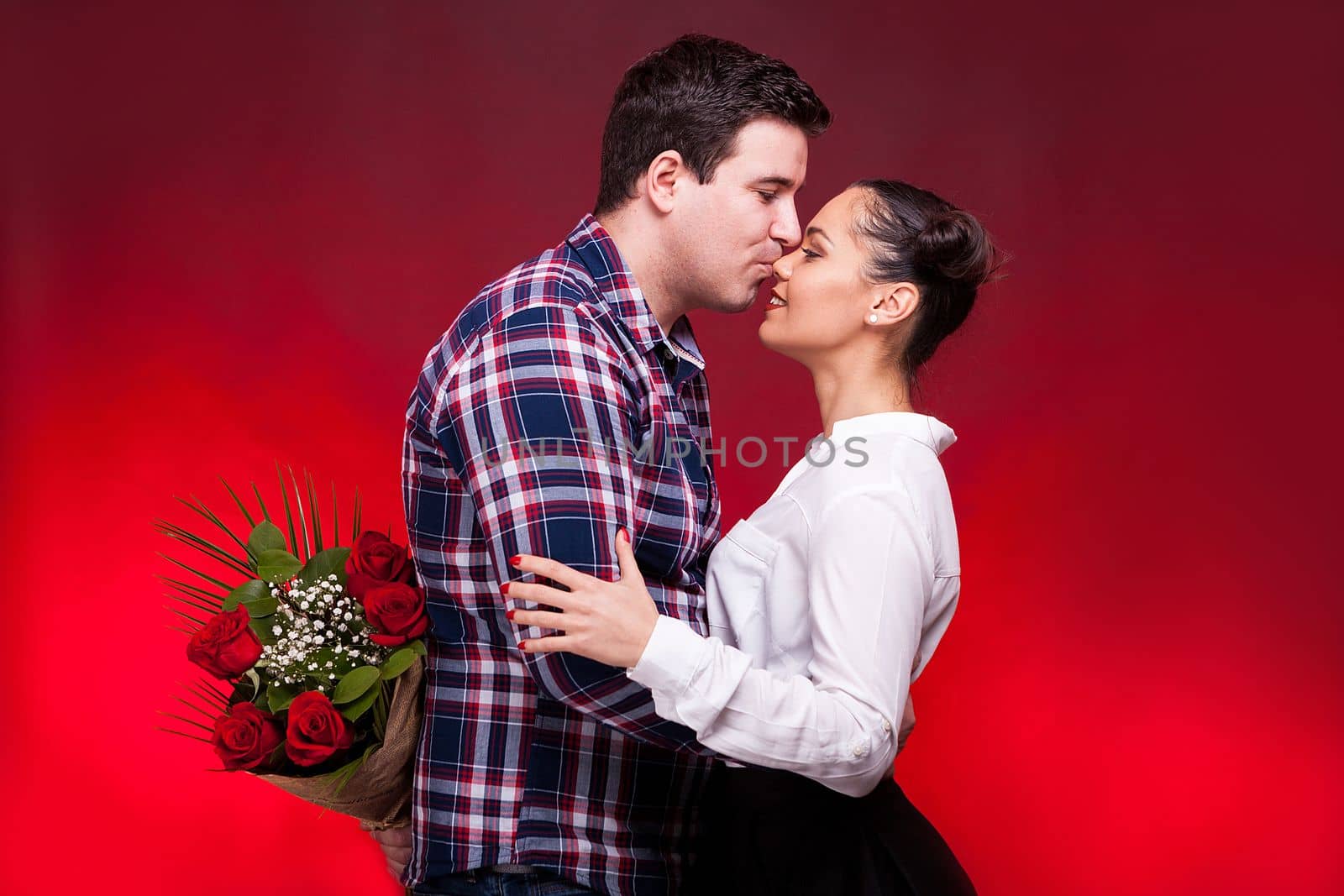 Man with a roses bouquet at his back kissing a beautiful woman on the nose. Red background and studio photo