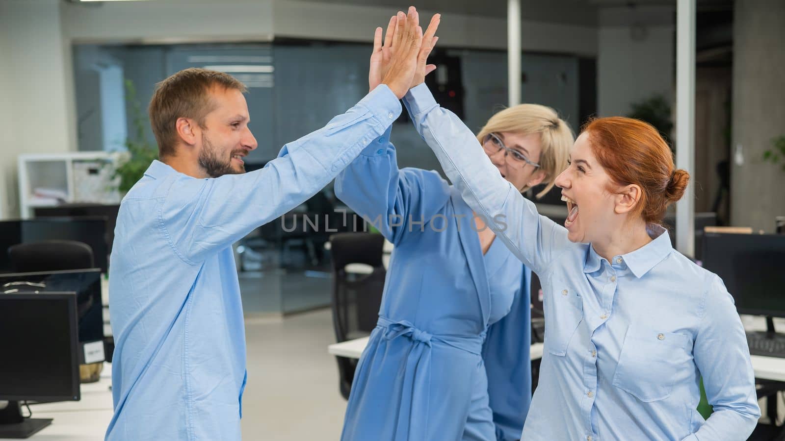 Colleagues give a high five in the office. Red-haired woman, blonde and bearded man in a denim shirt in the office