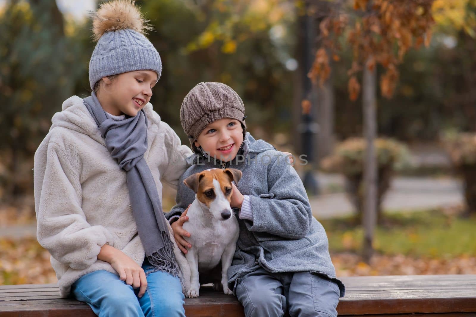 Brother and sister sit in an embrace with a dog on a bench for a walk in the autumn park. Boy, girl and jack russell terrier