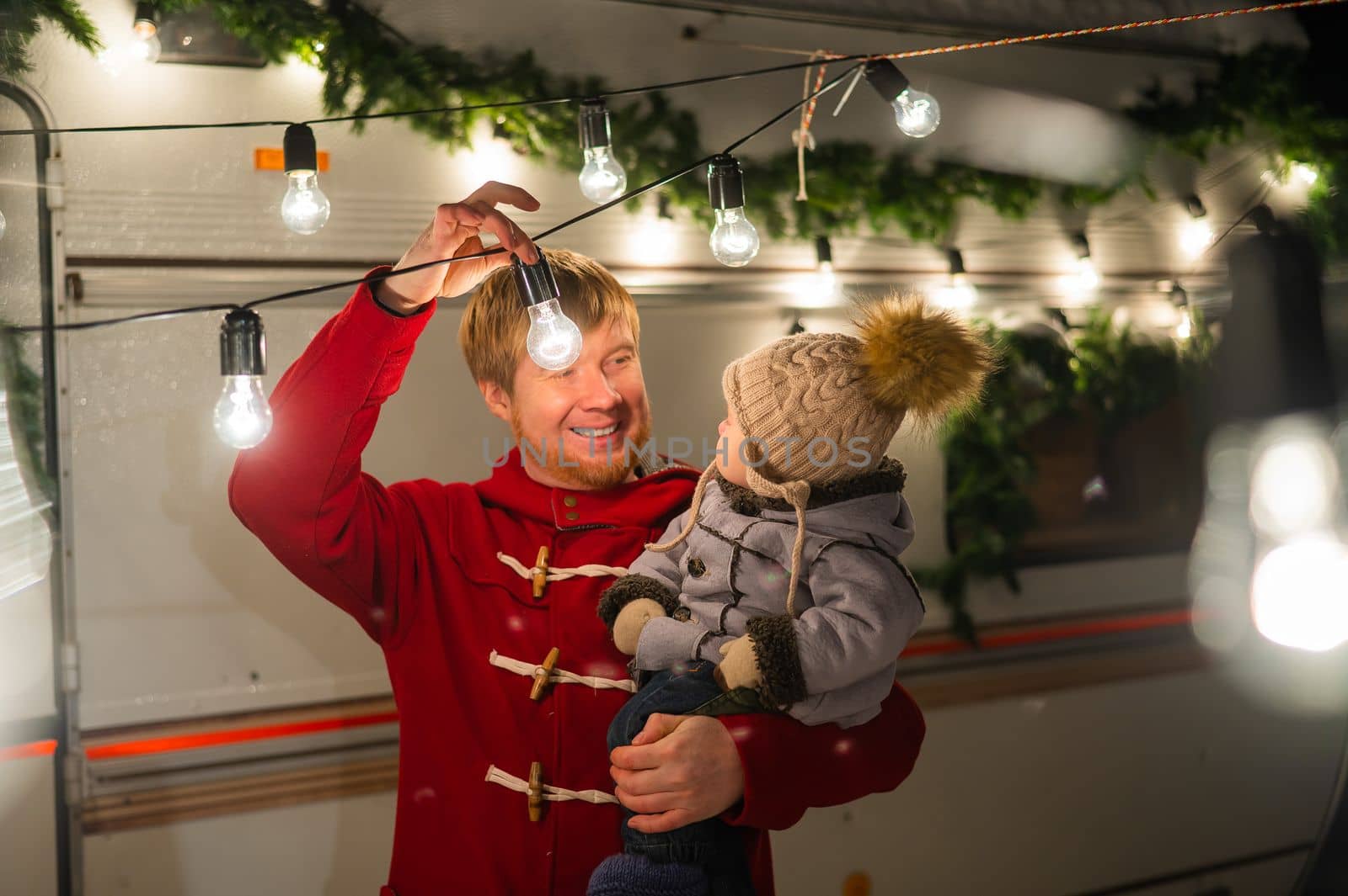 Caucasian red-haired man with a boy in his arms at the mobile home. Father and son celebrate Christmas on a trip