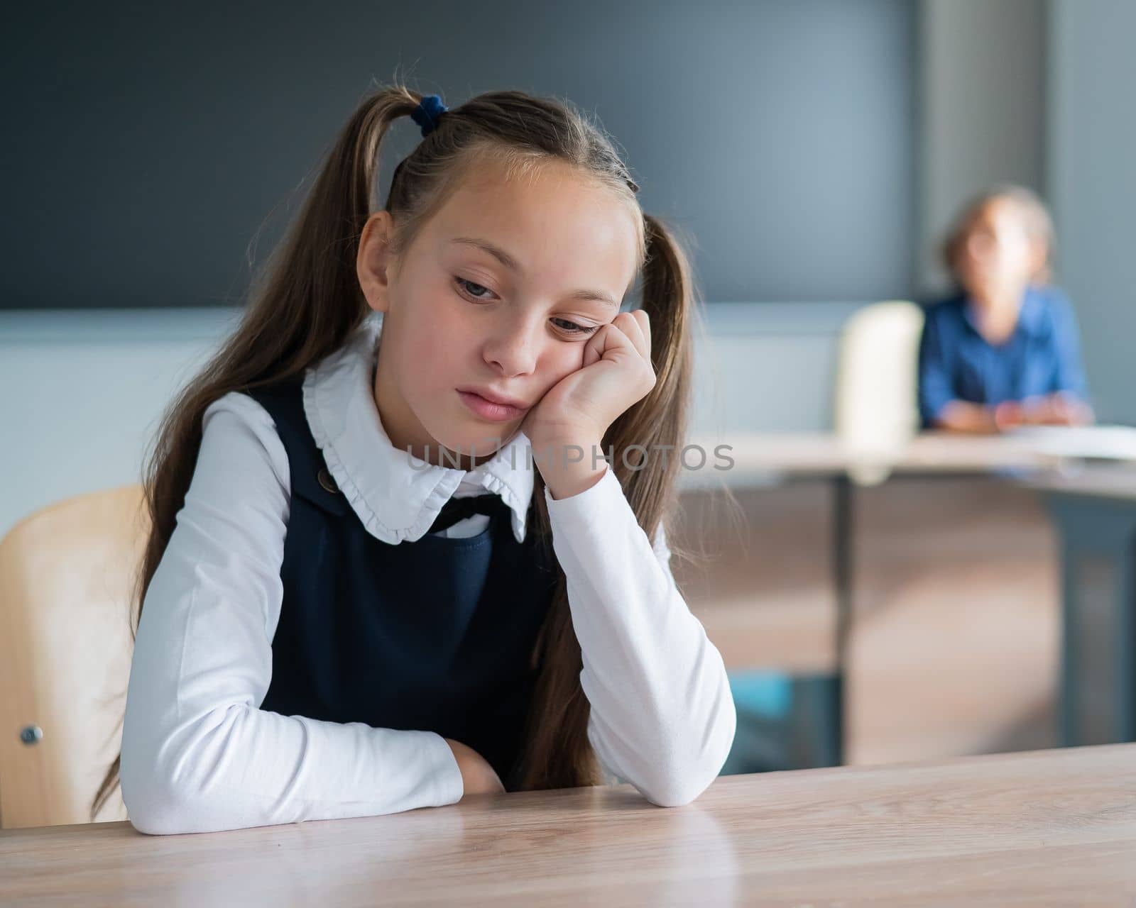 Little caucasian girl is bored at the lesson at school. The schoolgirl is sitting at her desk and the teacher is sitting in the background