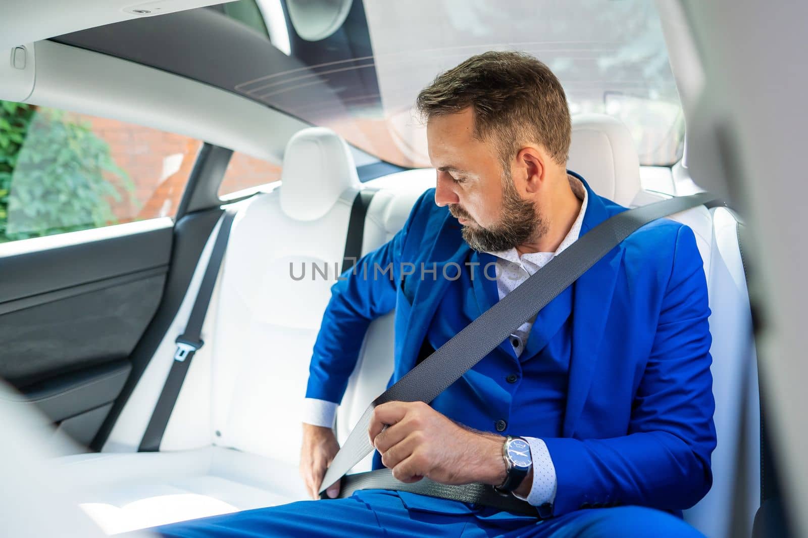 A caucasian man in a blue suit fastens his seat belt in the back seat of a car. Business class passenger