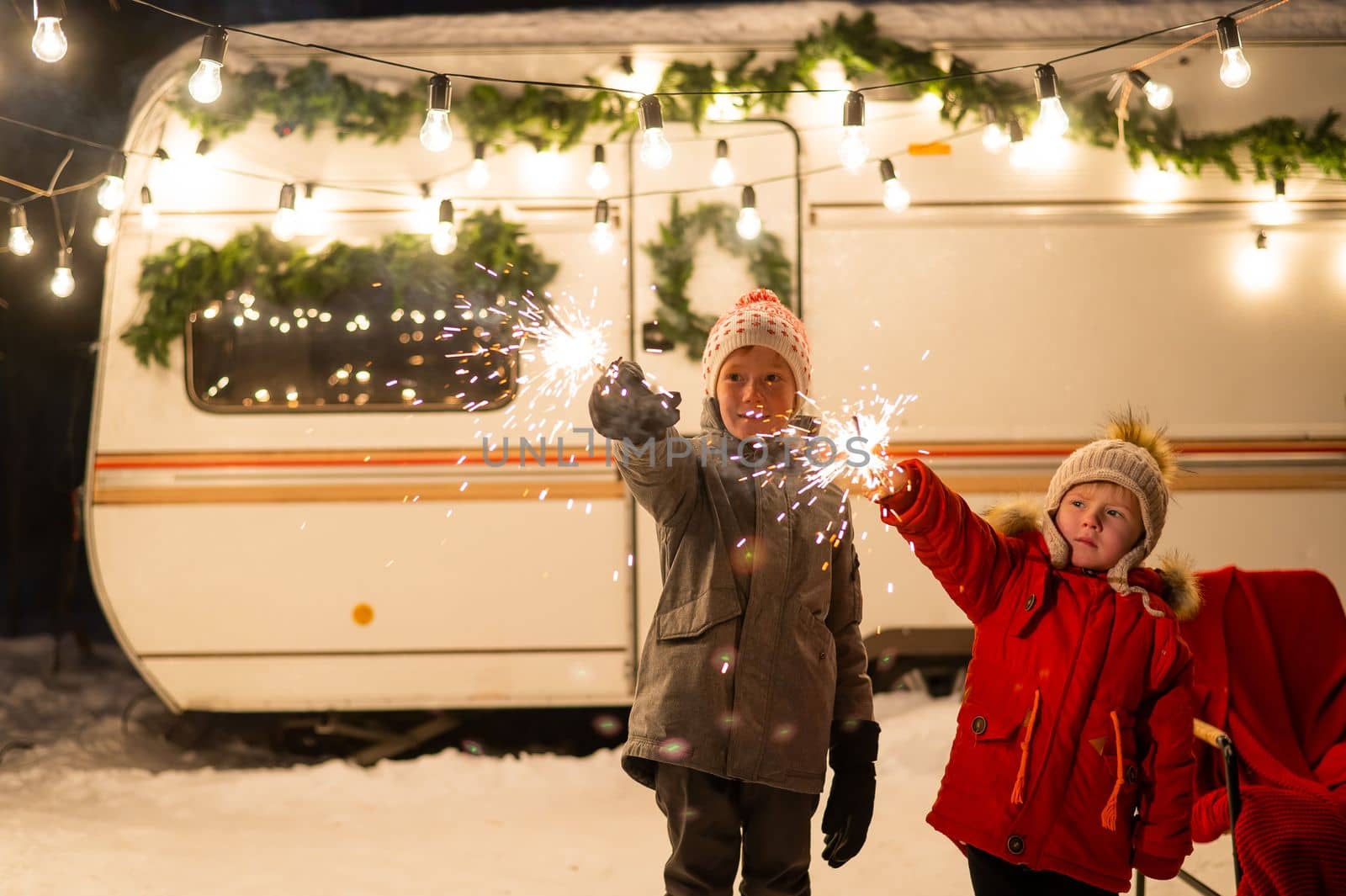 Caucasian red-haired boys hold sparklers by the trailer. Two brothers are celebrating Christmas on a trip