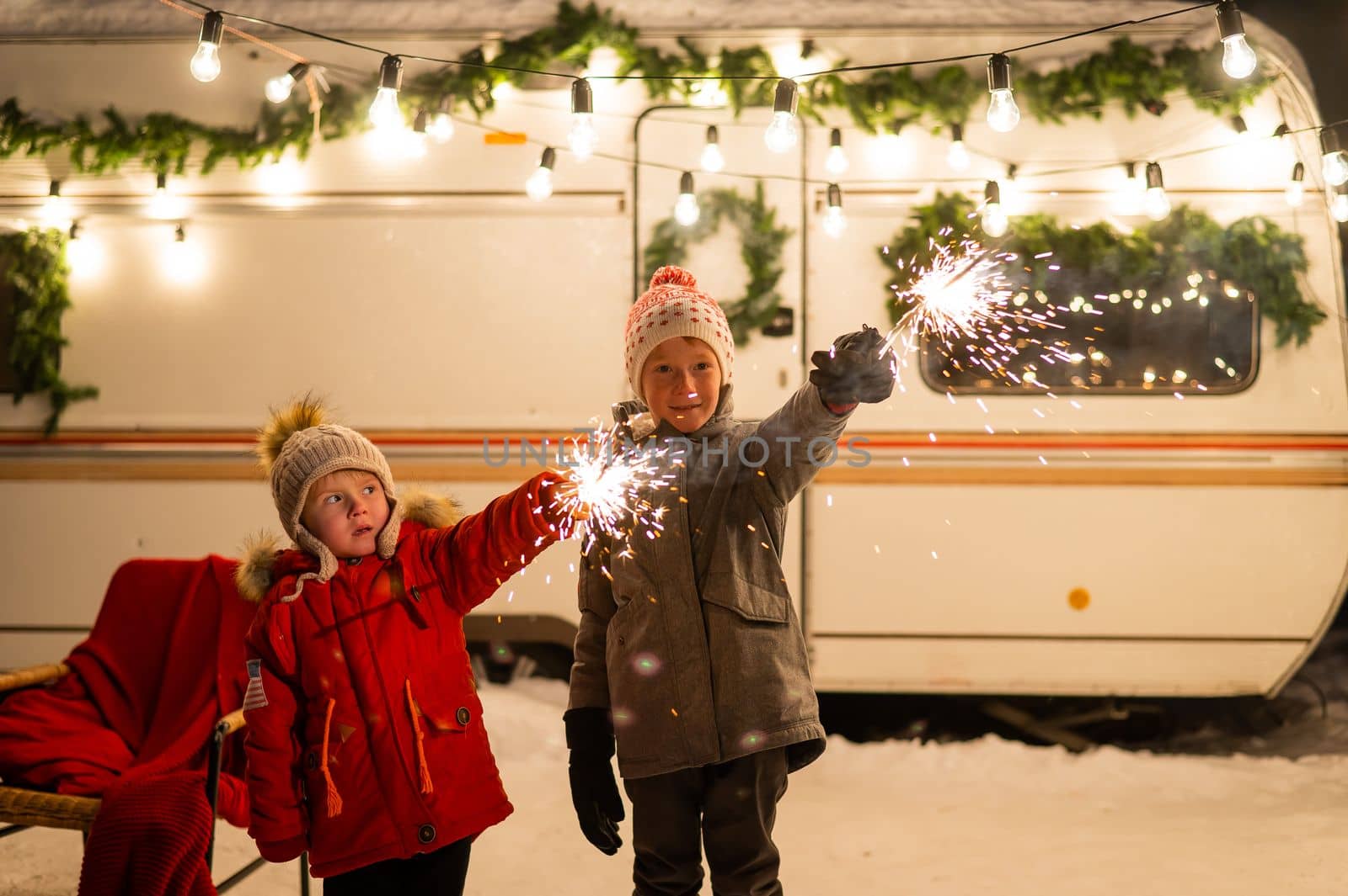 Caucasian red-haired boys hold sparklers by the trailer. Two brothers are celebrating Christmas on a trip