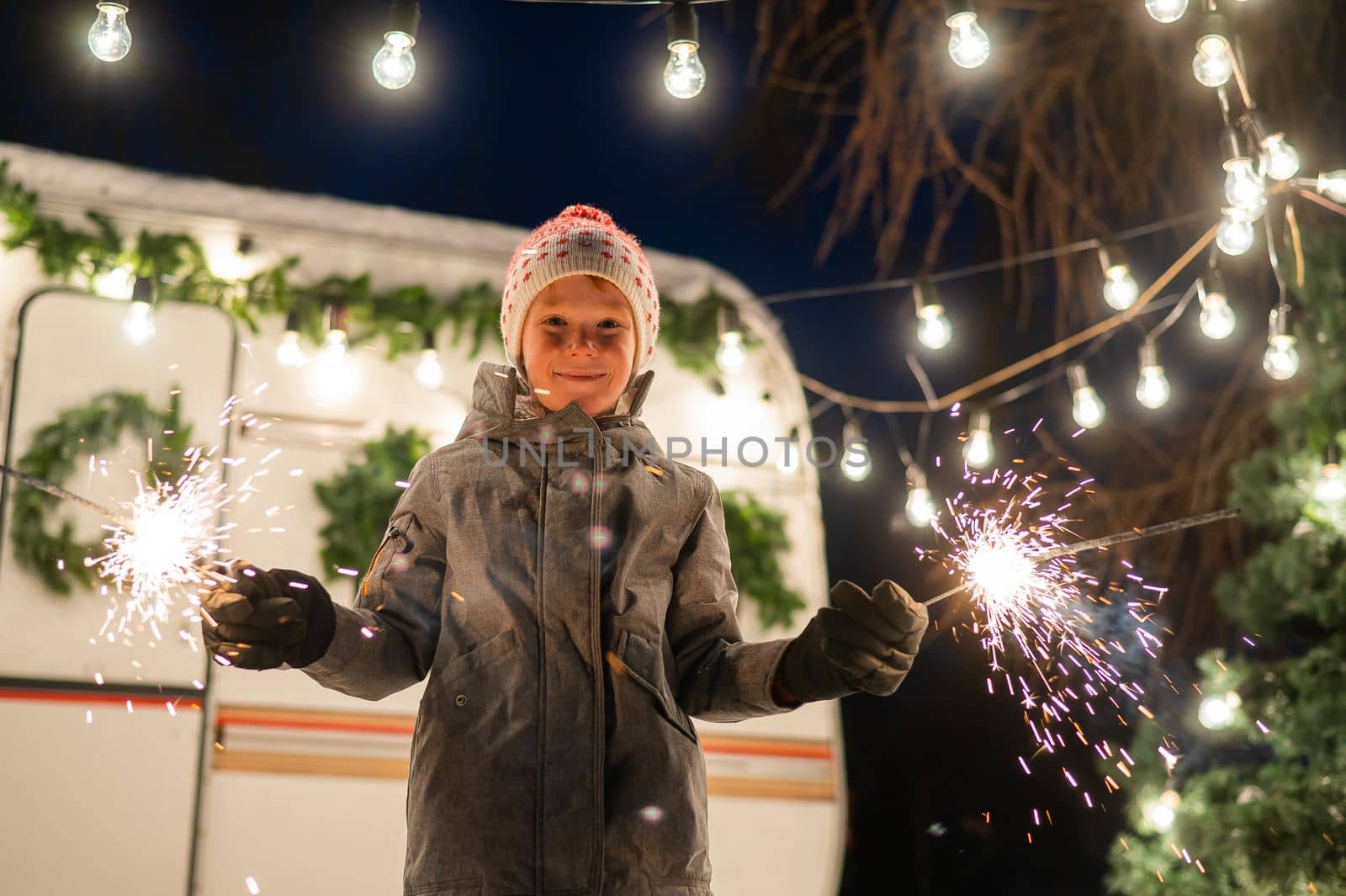 Caucasian red-haired boy holding sparklers by the trailer. Schoolboy celebrates Christmas on a trip