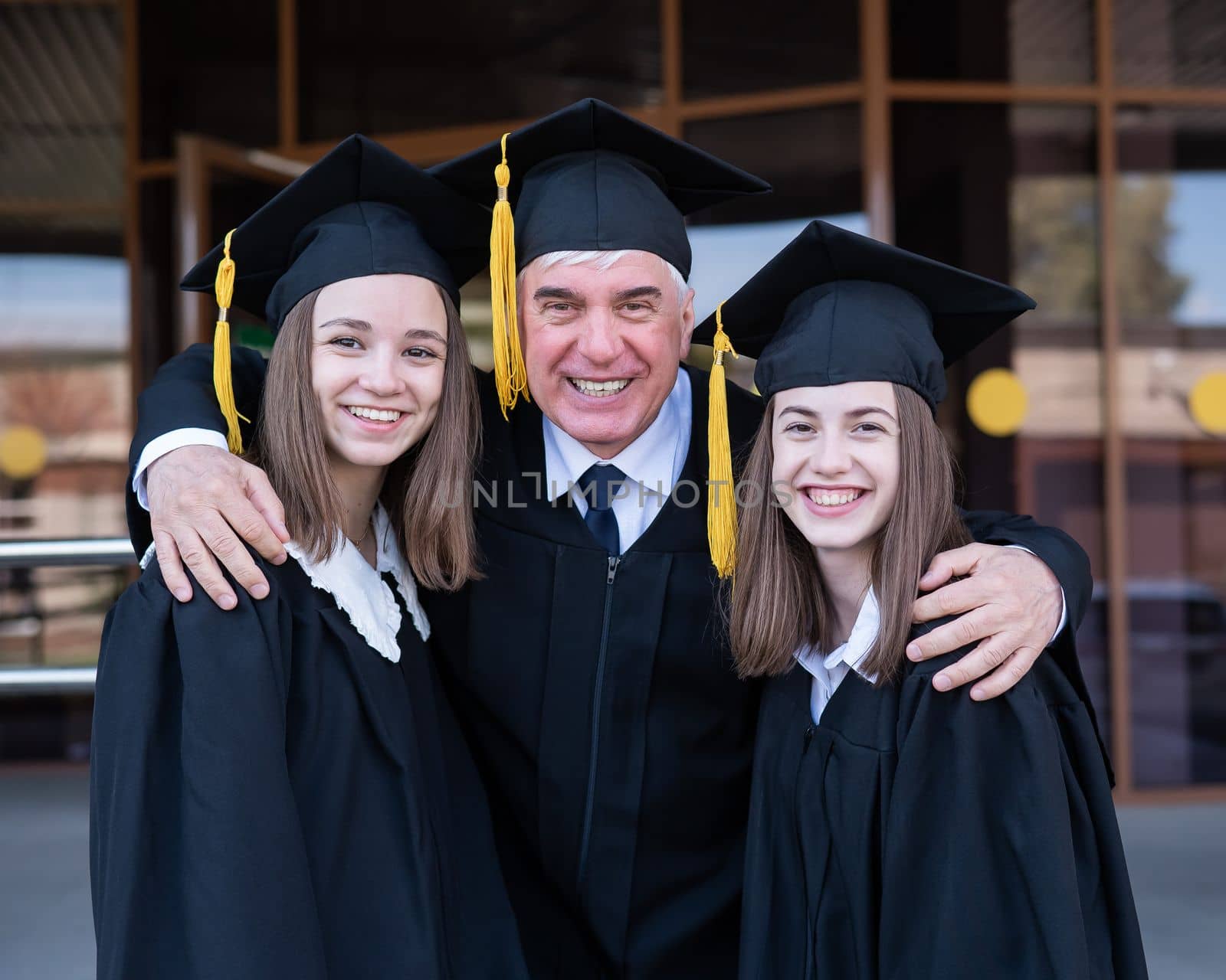 An elderly man hugs young girls. Classmates in graduation gowns