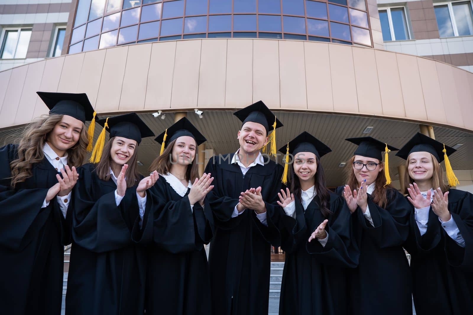 Seven graduates in robes stand in a row and clap outdoors