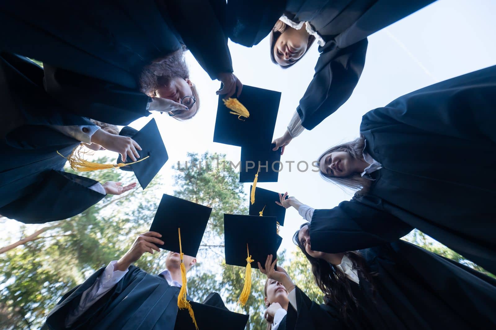 Classmates in graduation gowns toss their hats outdoors. Bottom view