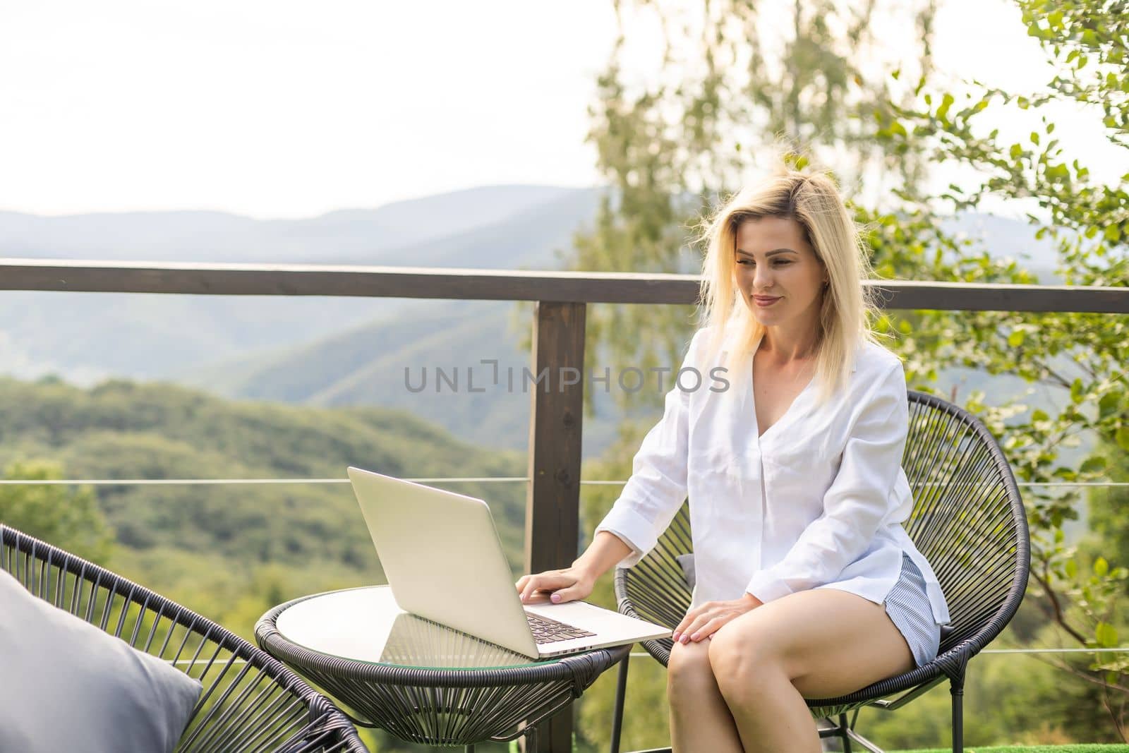 woman sitting on hotel balcony with laptop computer while working remote on freelance. mountains view.