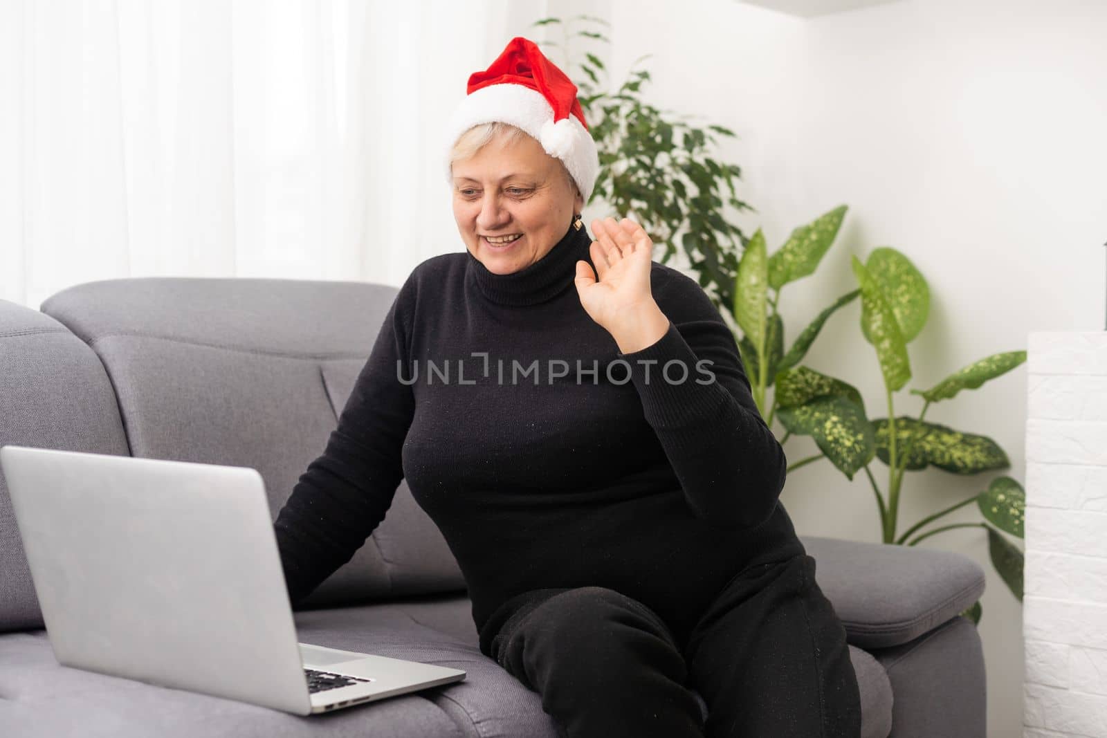 Smiling senior woman wearing Santa hat at Christmas time typing on laptop from home getting in touch with family or friends. by Andelov13