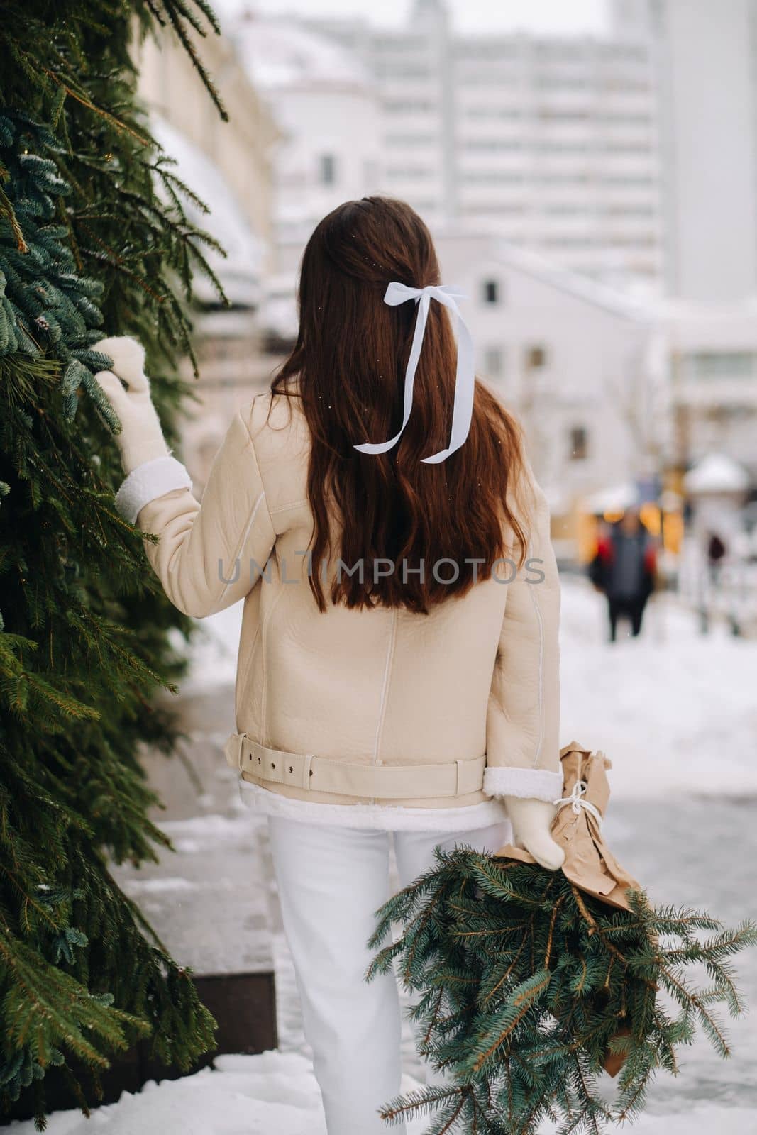 A girl with long hair stands with her back in the city with a bouquet of fir branches. Snowy winter by Lobachad