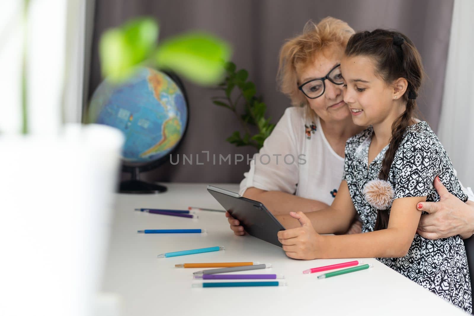 Happy mature grandmother with adorable little granddaughter using tablet at home together, excited middle aged woman and cute kid looking at device screen.