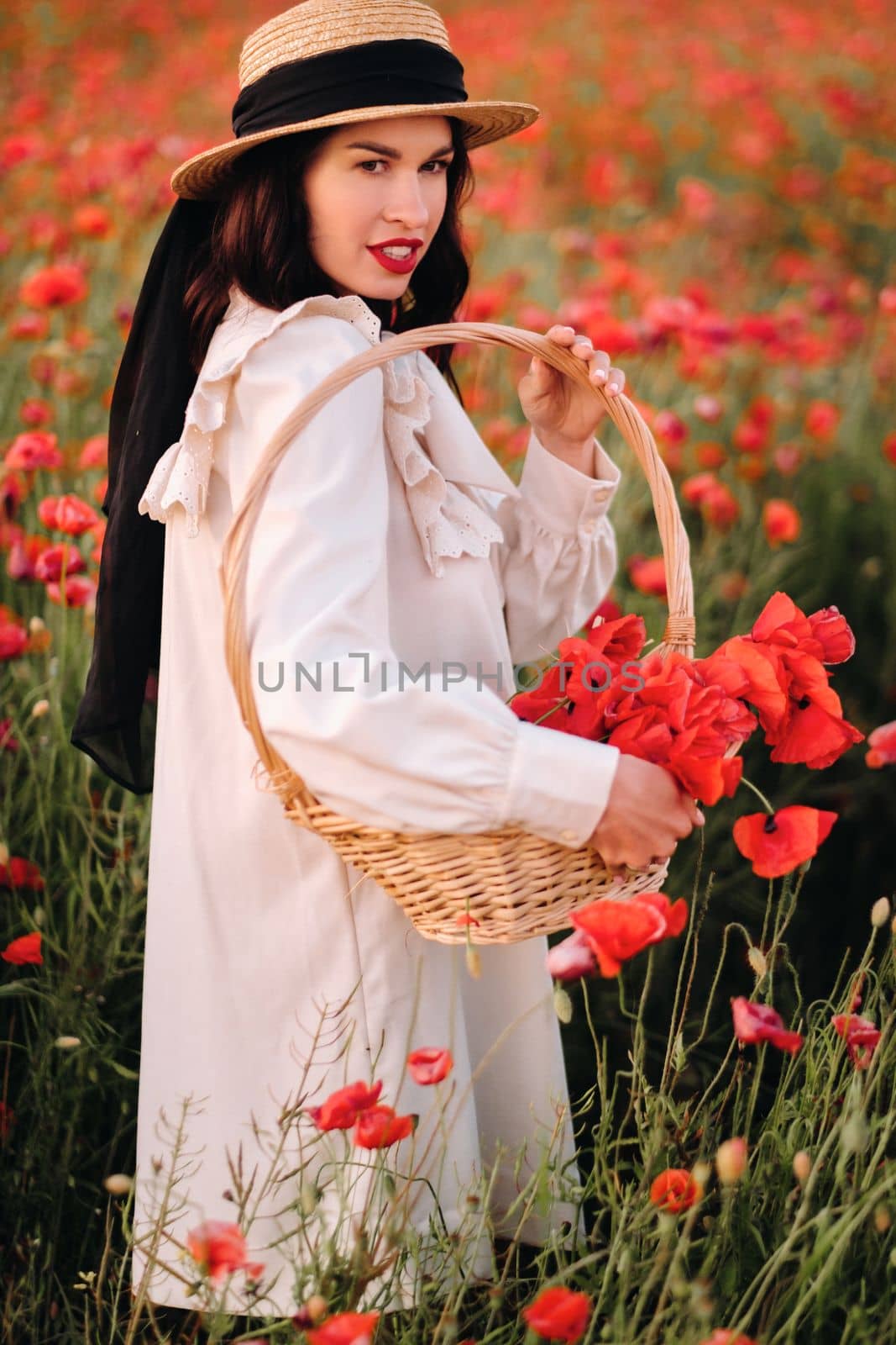 A girl in a white dress and with a basket of poppies walks through a poppy field.