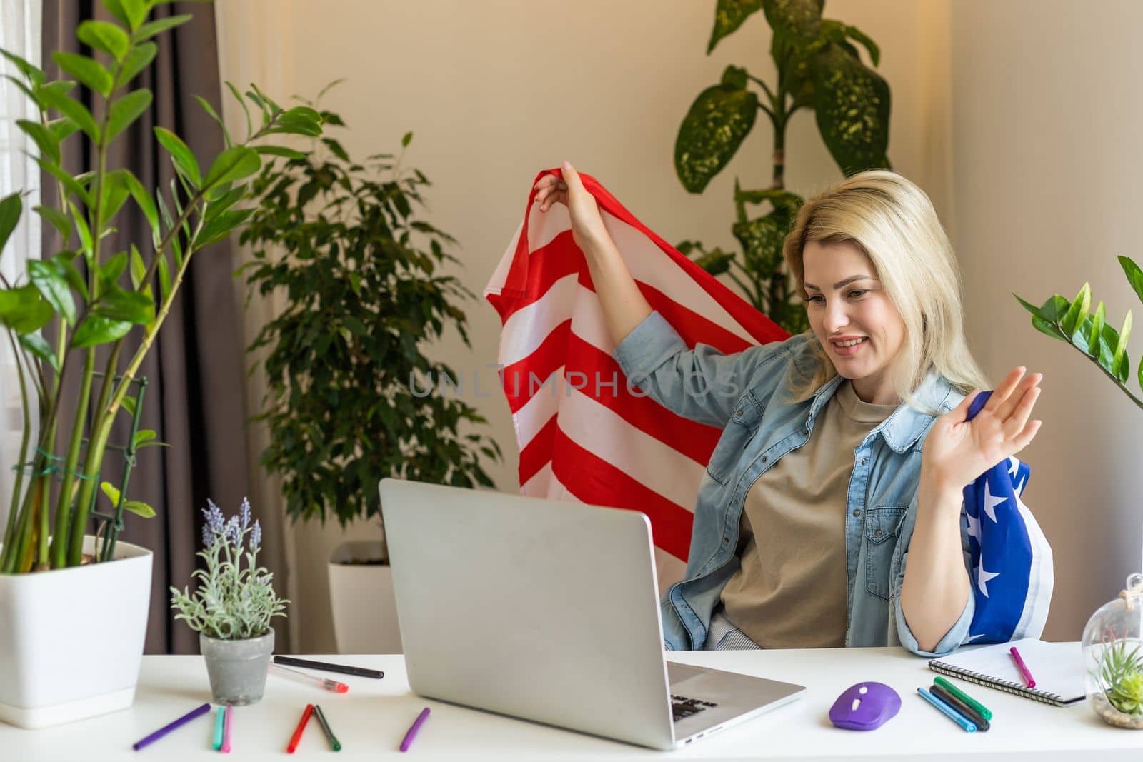 Happy woman employee sitting wrapped in USA flag, shouting for joy in office workplace, celebrating labor day or US Independence day. Indoor studio studio shot isolated on yellow background.