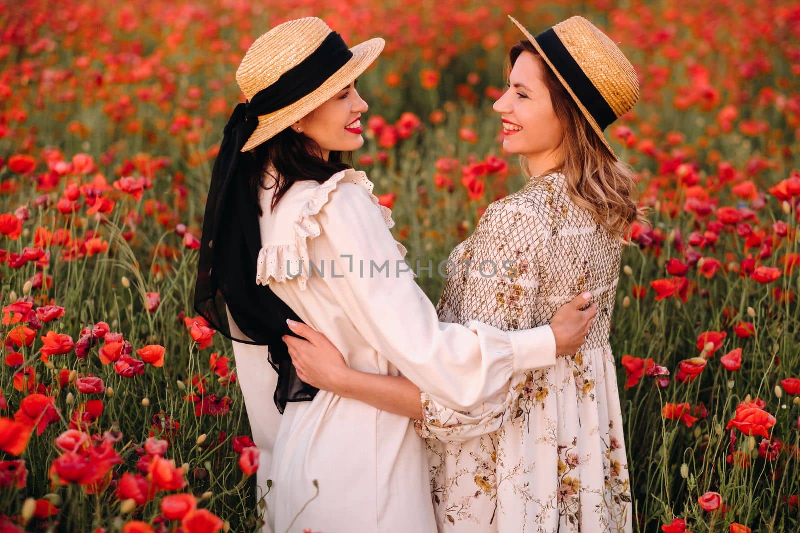 Two girlfriends in dresses and a hat in a poppy field in summer at sunset.