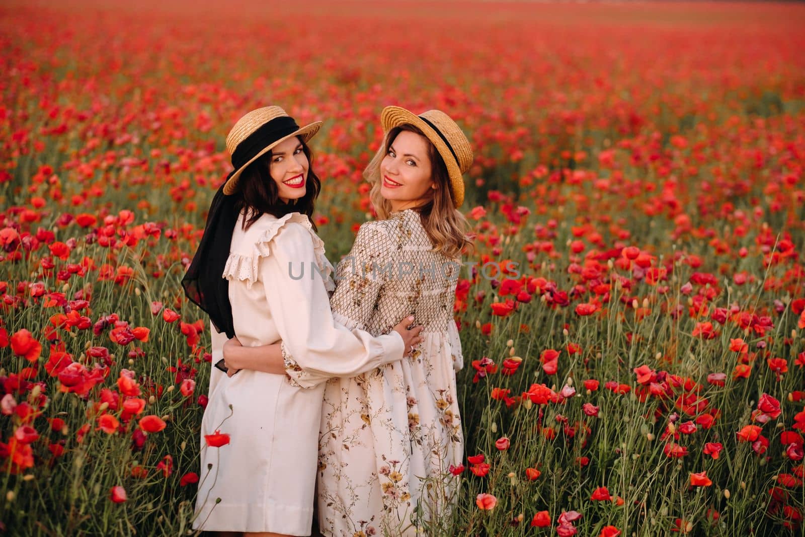 Two girlfriends in dresses and a hat in a poppy field in summer at sunset.