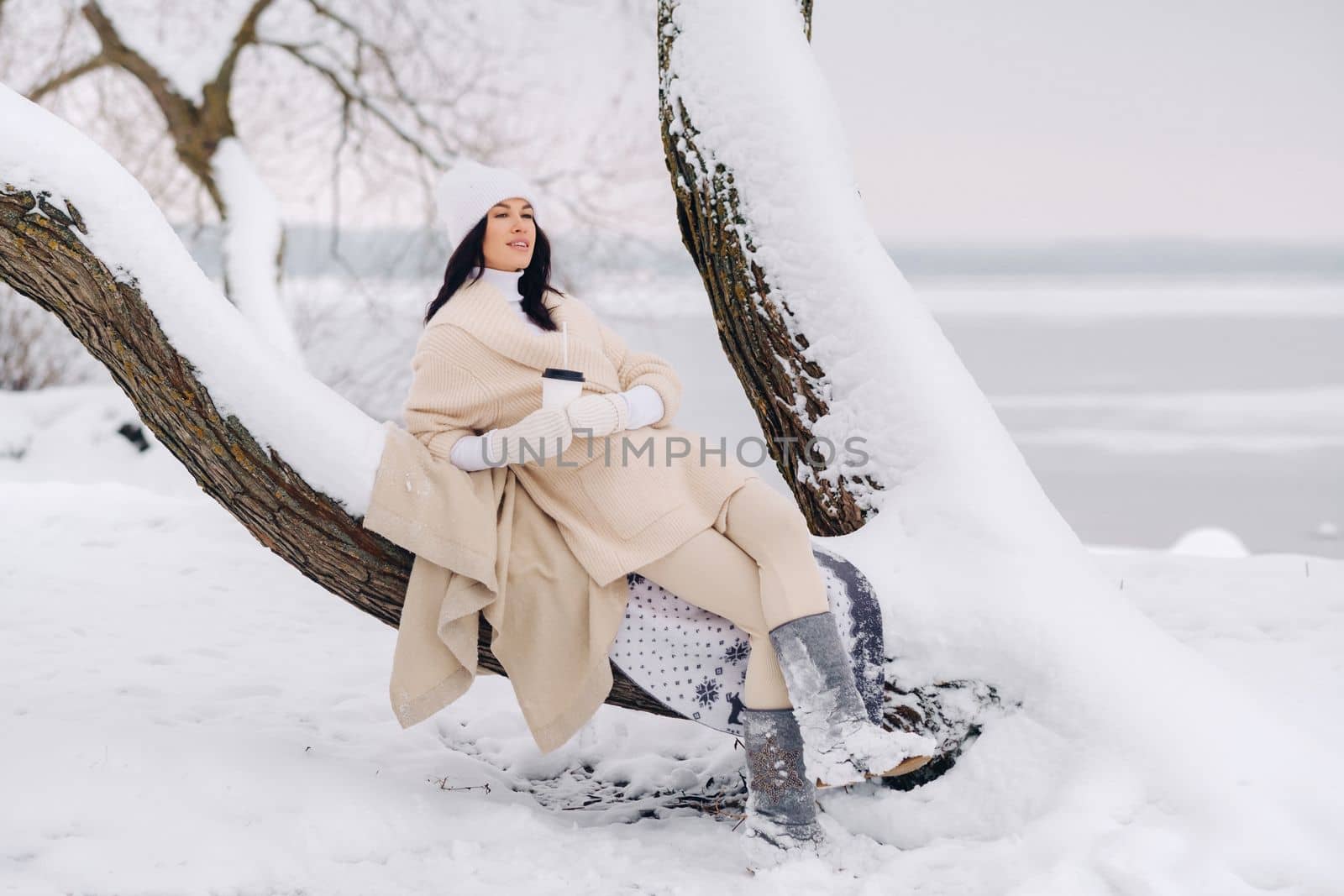 A beautiful girl with a beige cardigan and a white hat enjoying drinking tea in a snowy winter forest near a lake.