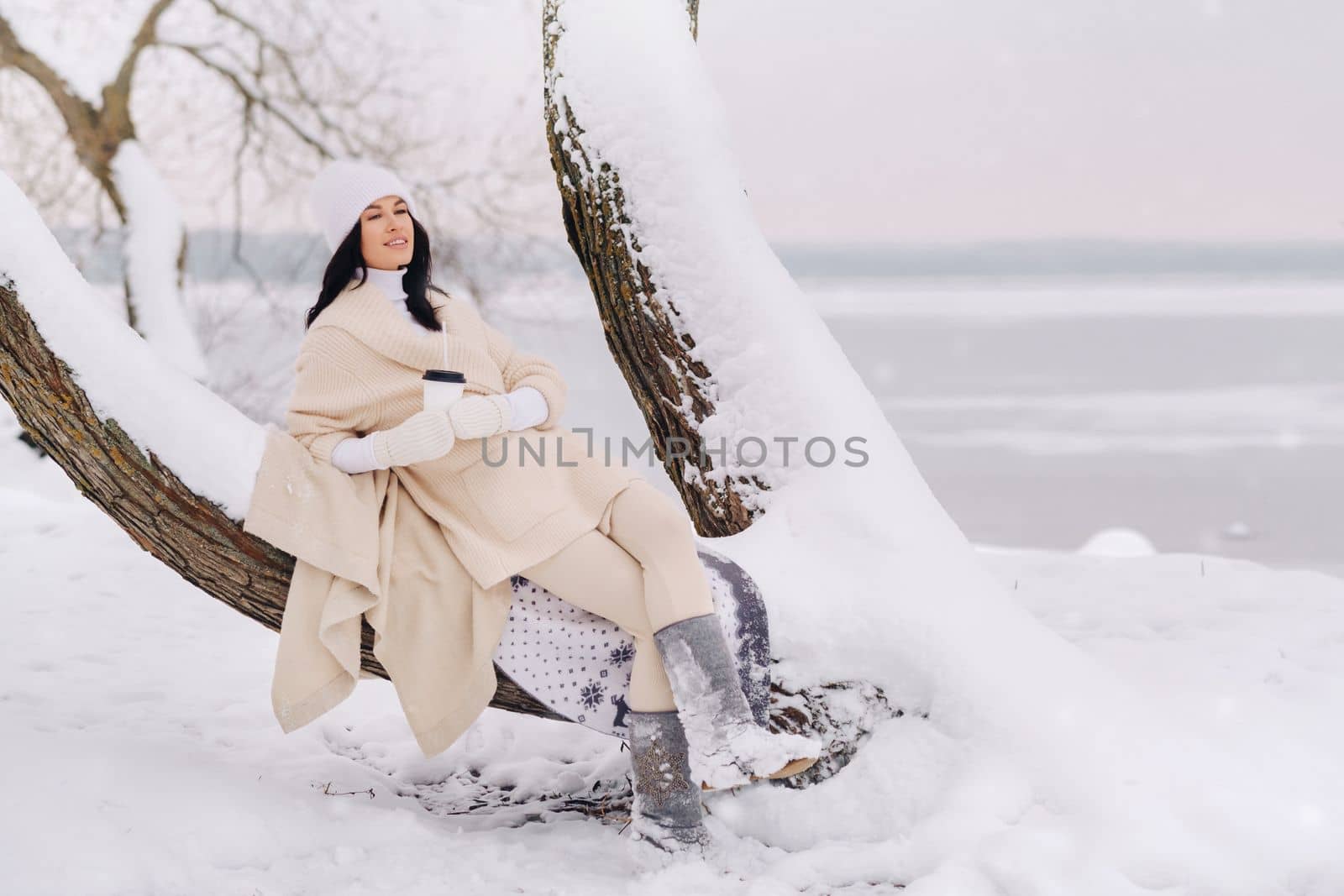 A beautiful girl with a beige cardigan and a white hat enjoying drinking tea in a snowy winter forest near a lake.