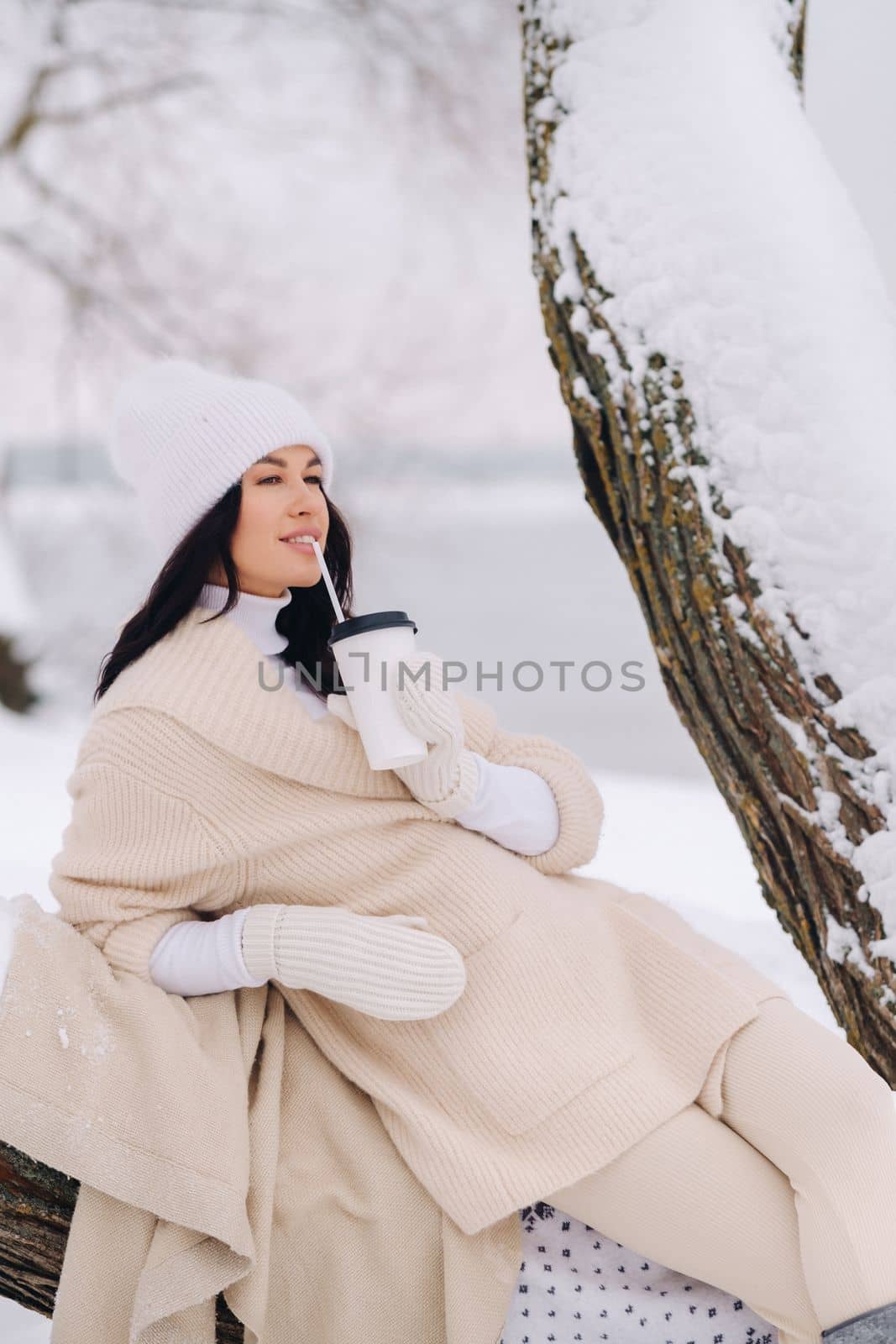 A beautiful girl with a beige cardigan and a white hat enjoying drinking tea in a snowy winter forest near a lake by Lobachad