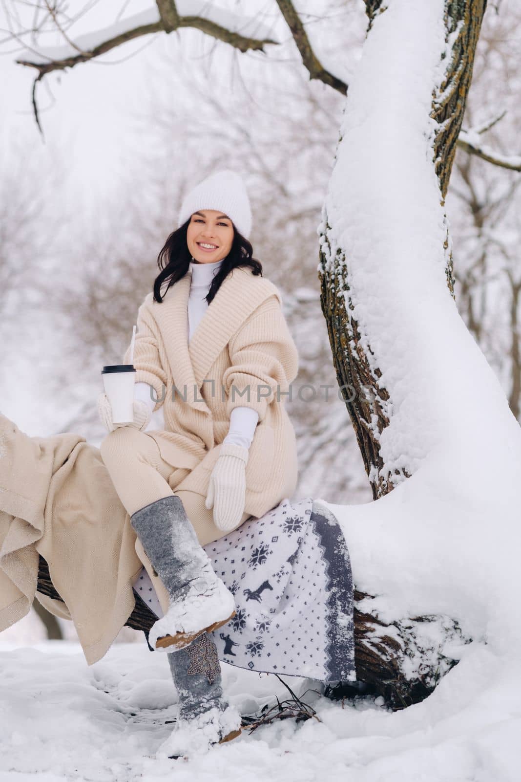 A beautiful girl with a beige cardigan and a white hat enjoying drinking tea in a snowy winter forest near a lake by Lobachad