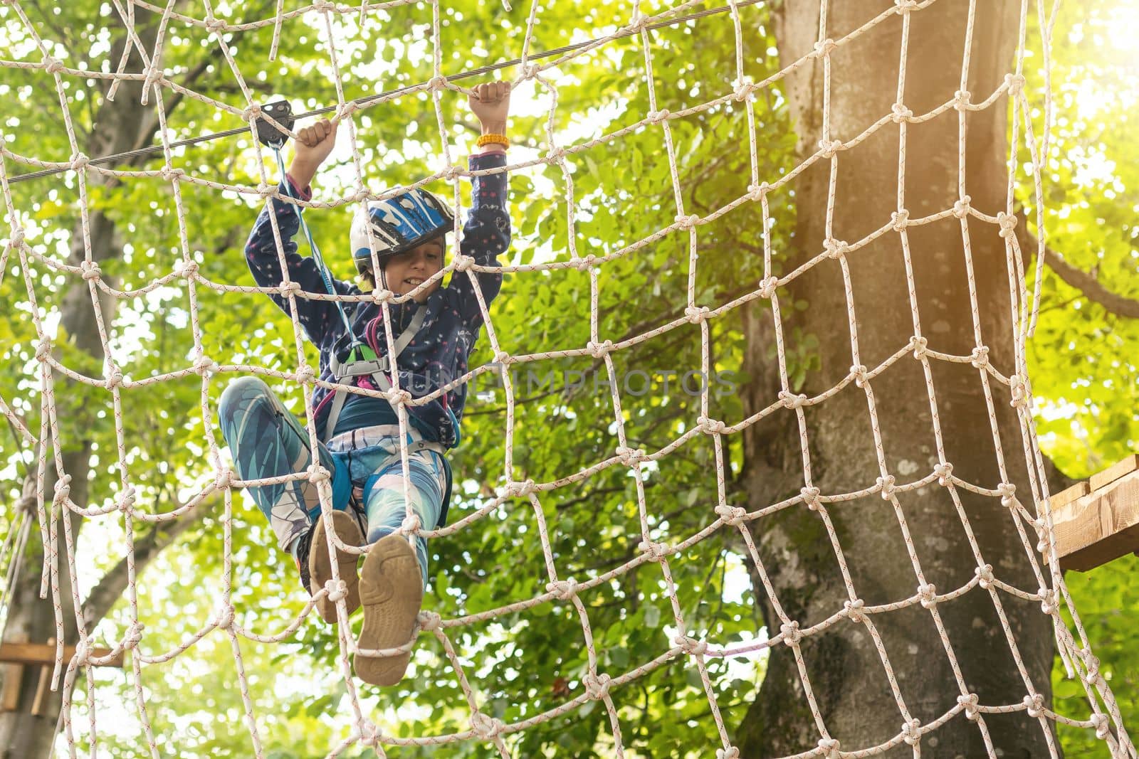 Little girl preschooler wearing full climbing harness having fun time in the rope park using carabiner and other safety equipment. Summer camp activity for kids. Adventure park in the forest.