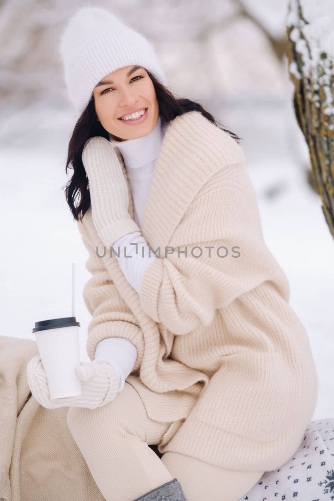 A beautiful girl with a beige cardigan and a white hat enjoying drinking tea in a snowy winter forest near a lake by Lobachad