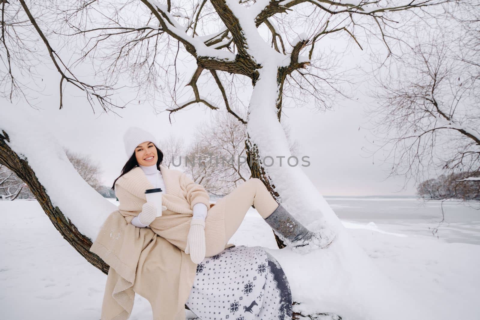 A beautiful girl with a beige cardigan and a white hat enjoying drinking tea in a snowy winter forest near a lake.