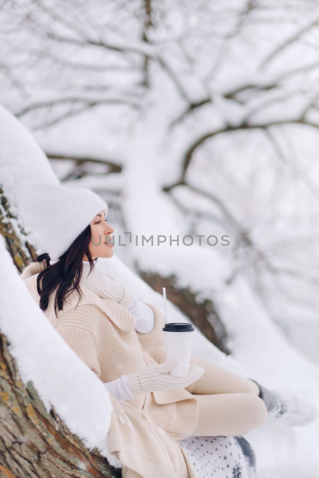 A beautiful girl with a beige cardigan and a white hat enjoying drinking tea in a snowy winter forest near a lake by Lobachad