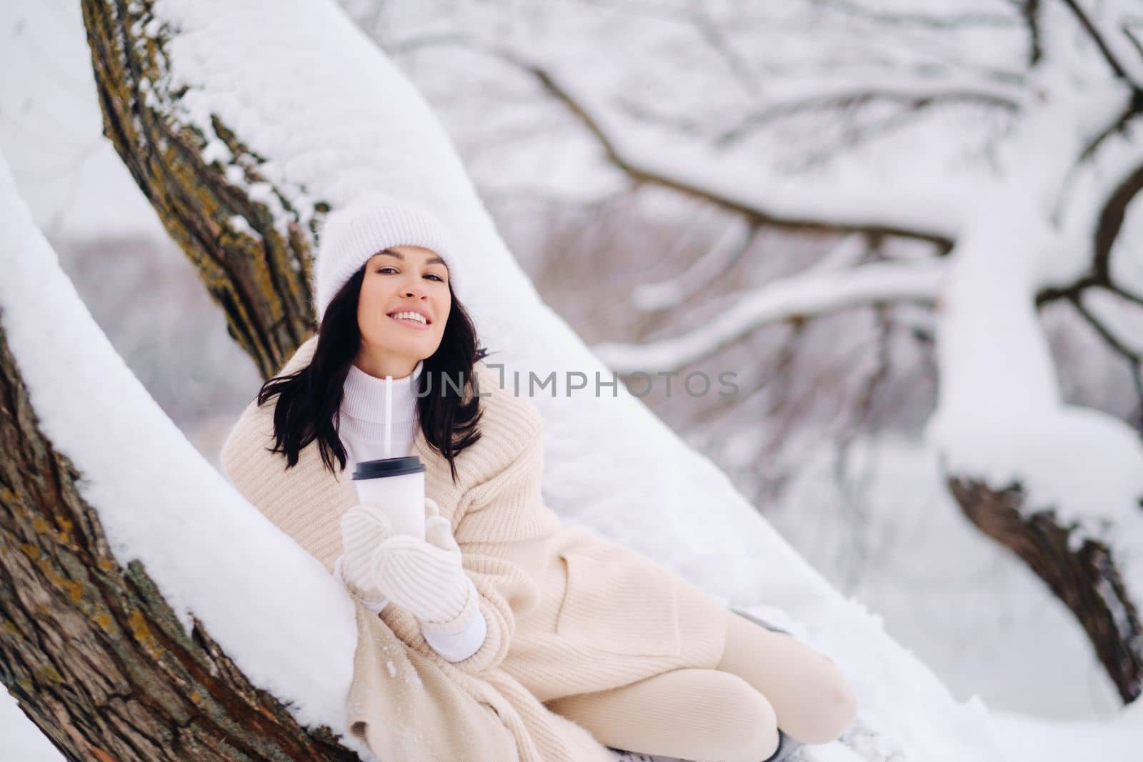 A beautiful girl with a beige cardigan and a white hat enjoying drinking tea in a snowy winter forest near a lake.