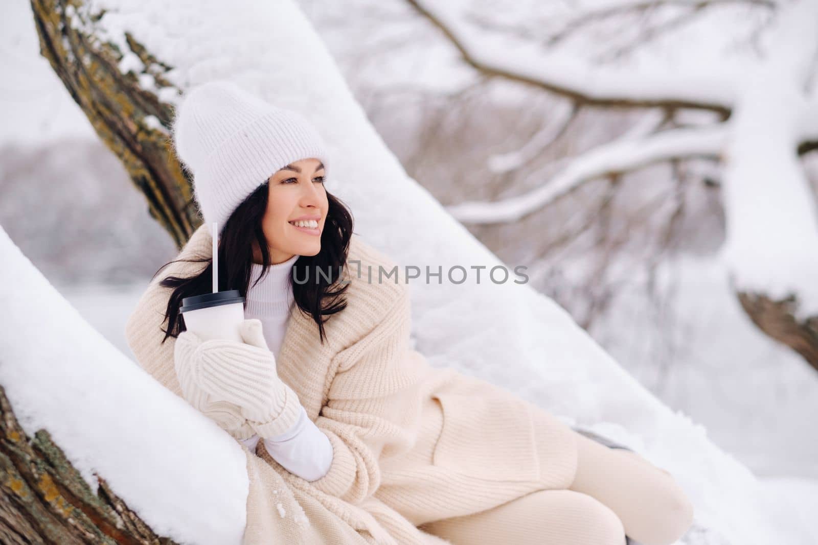 A beautiful girl with a beige cardigan and a white hat enjoying drinking tea in a snowy winter forest near a lake.