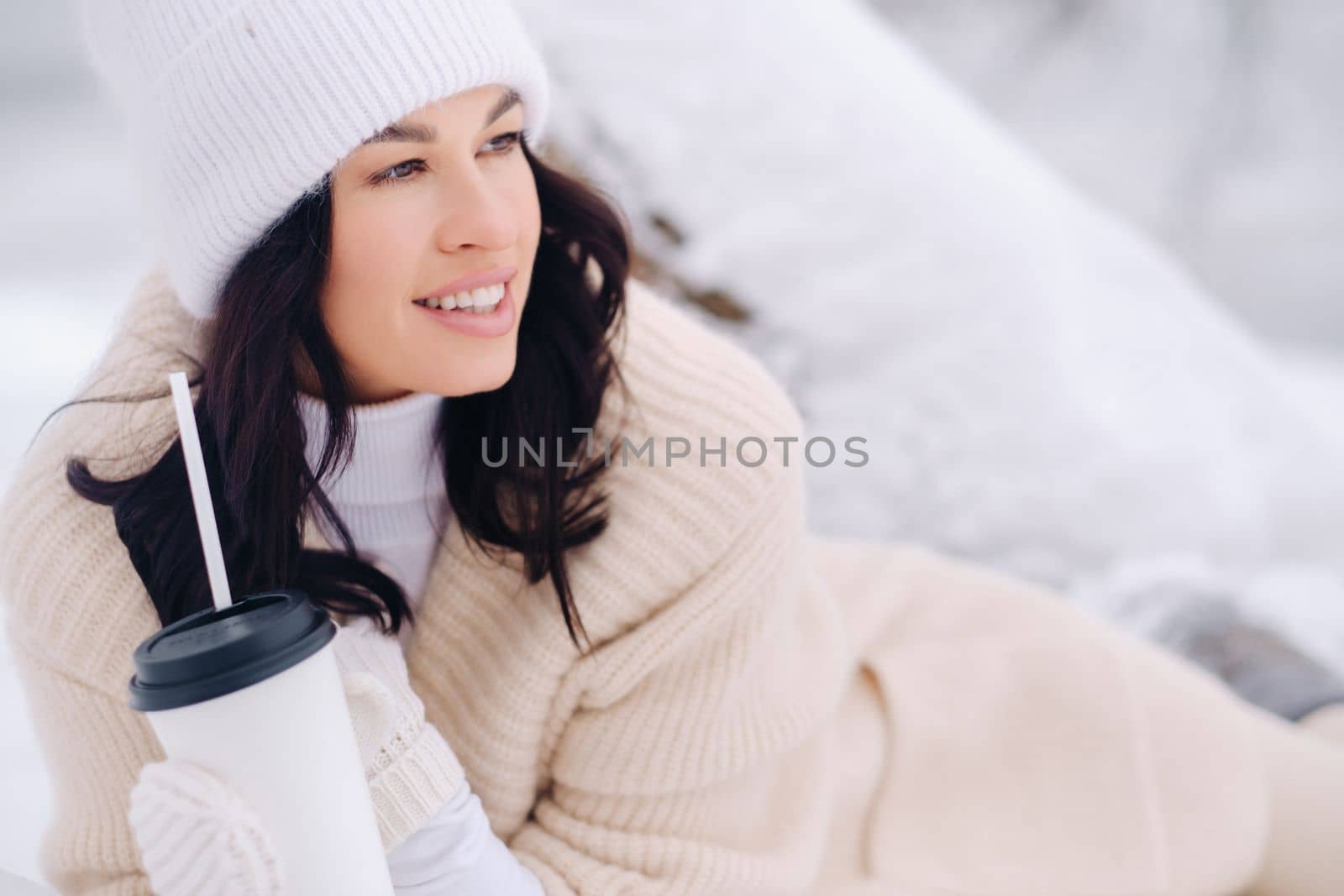 A beautiful girl with a beige cardigan and a white hat enjoying drinking tea in a snowy winter forest near a lake.