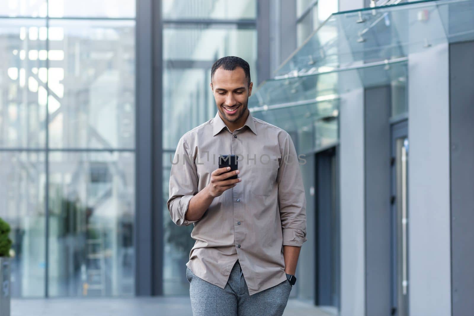 Latin american businessman in casual shirt walking near office building from outside, man smiling and using phone, freelancer typing message and browsing internet pages.