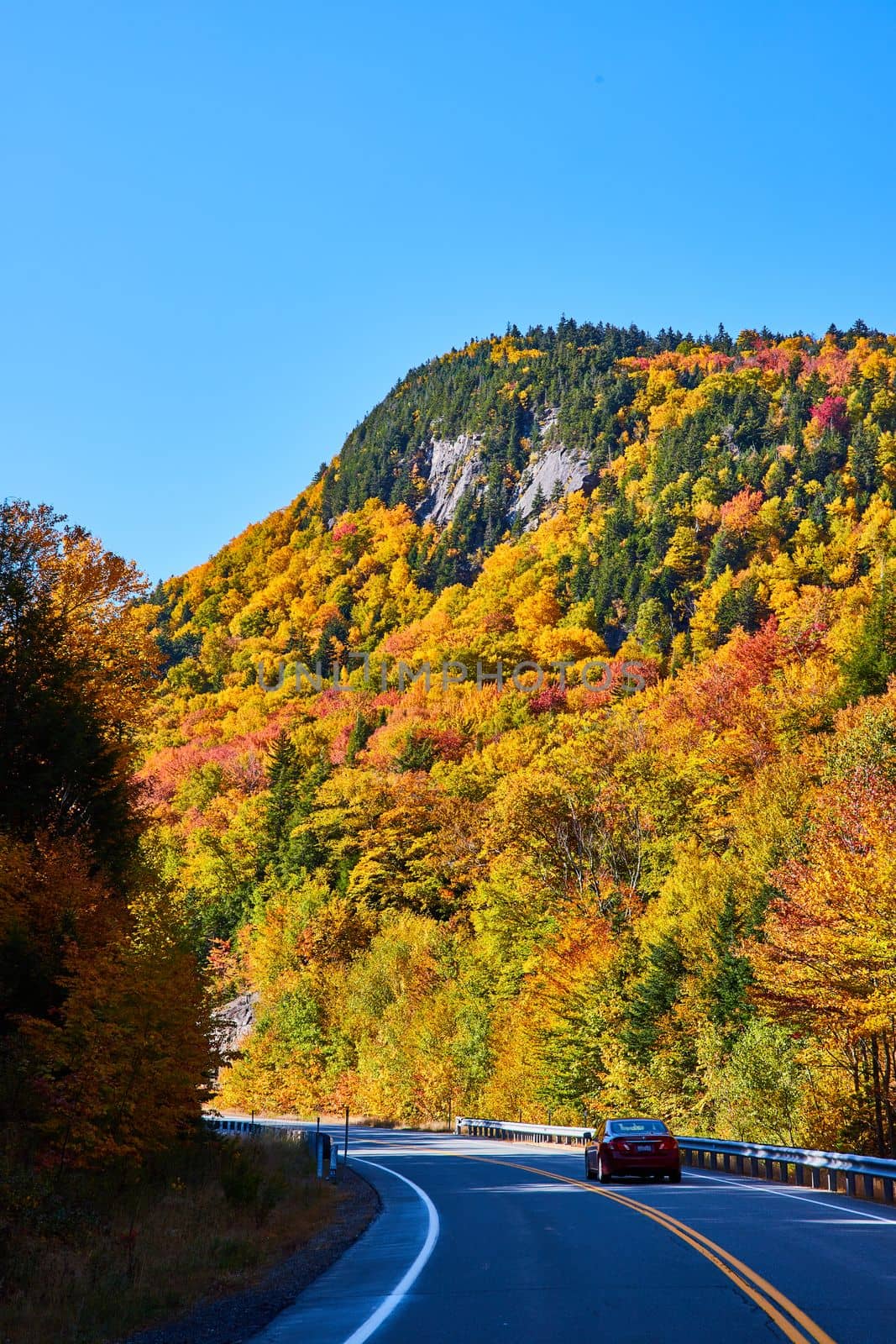 Image of Beautiful foliage covers rocky mountain viewed from road with car passing through