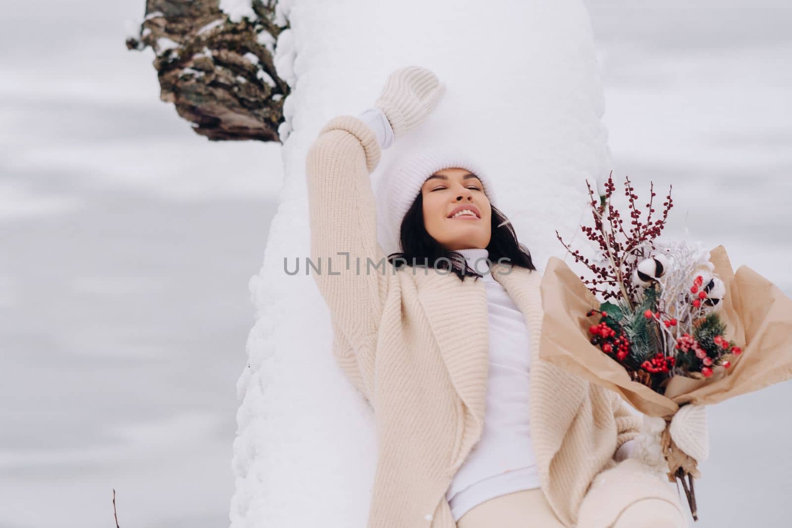 A beautiful girl in a beige cardigan and a white hat with flowers enjoys a snowy winter in nature.